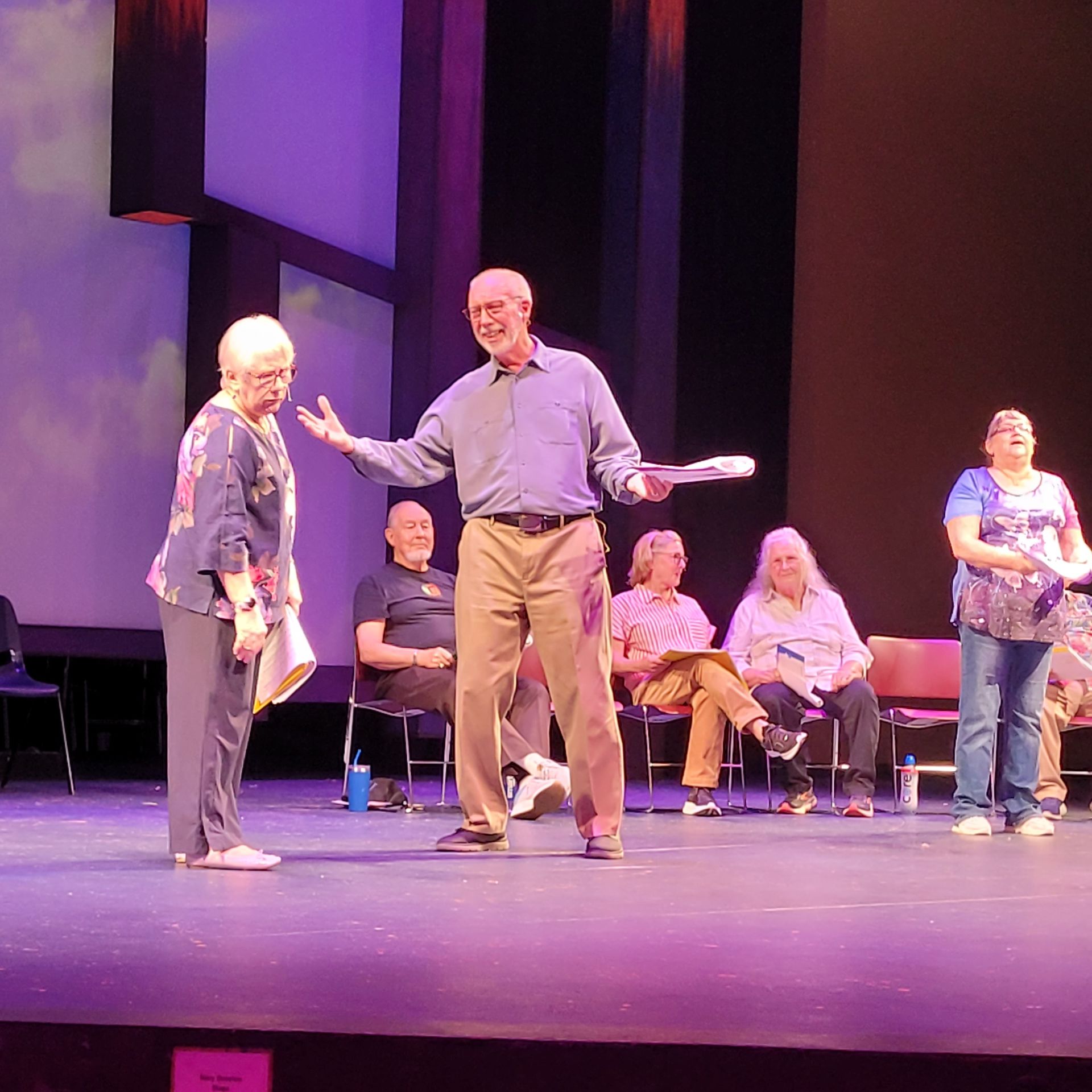 Onstage: Actors performing a play. Man gesturing, woman listening, seated audience watches. Purple lighting.