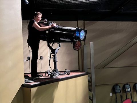 Man adjusts a large spotlight on a platform in a theater, looking at the camera.