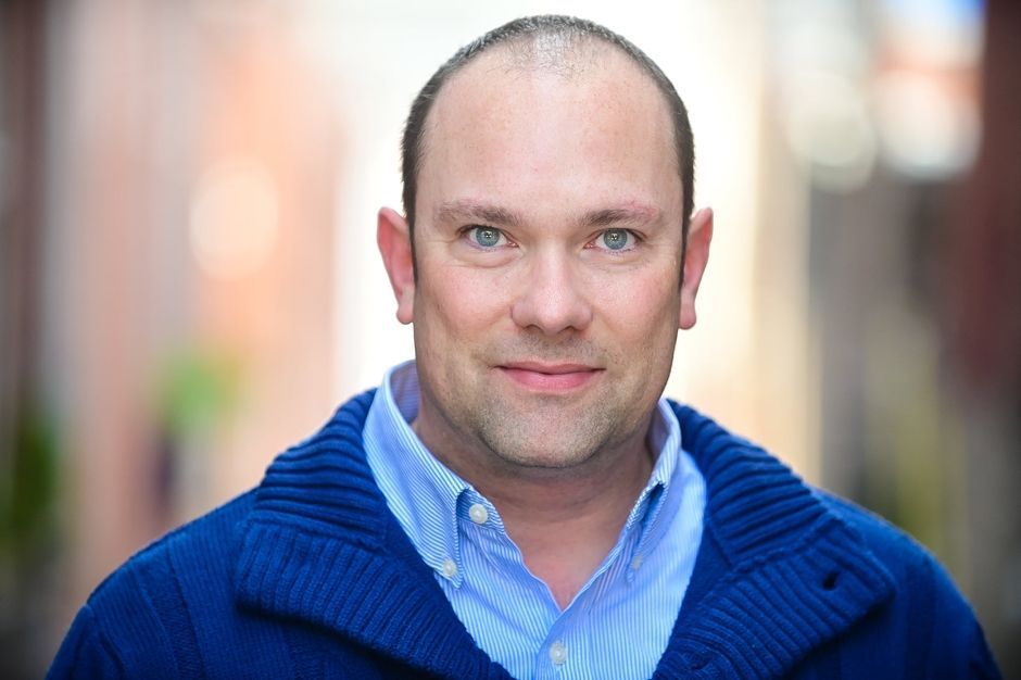 Smiling man in a blue sweater and shirt, standing outdoors with a blurred street background