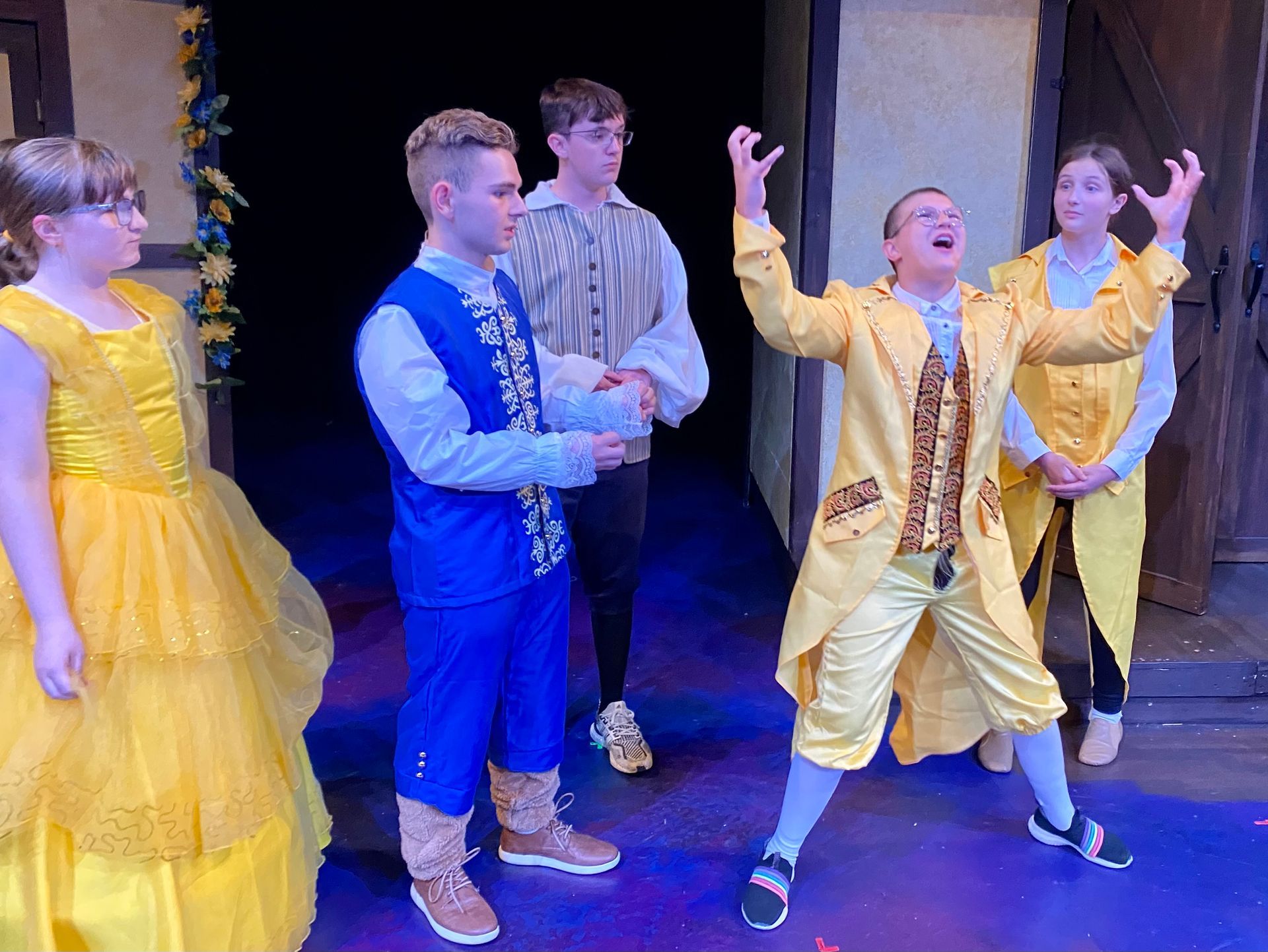 Theater production: Cast in costume on stage; yellow & blue outfits, excited actor in focus.