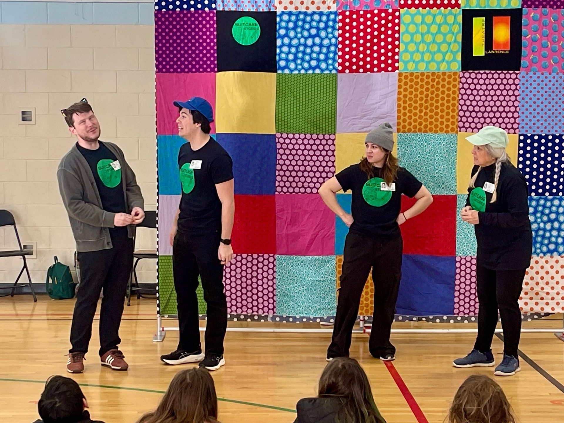 Four people on a stage in front of a colorful quilt backdrop; all wearing black shirts with a green circle logo.