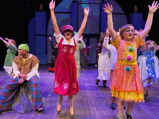 Children on a stage in colorful costumes, arms raised, celebrating. Theater backdrop in blue.