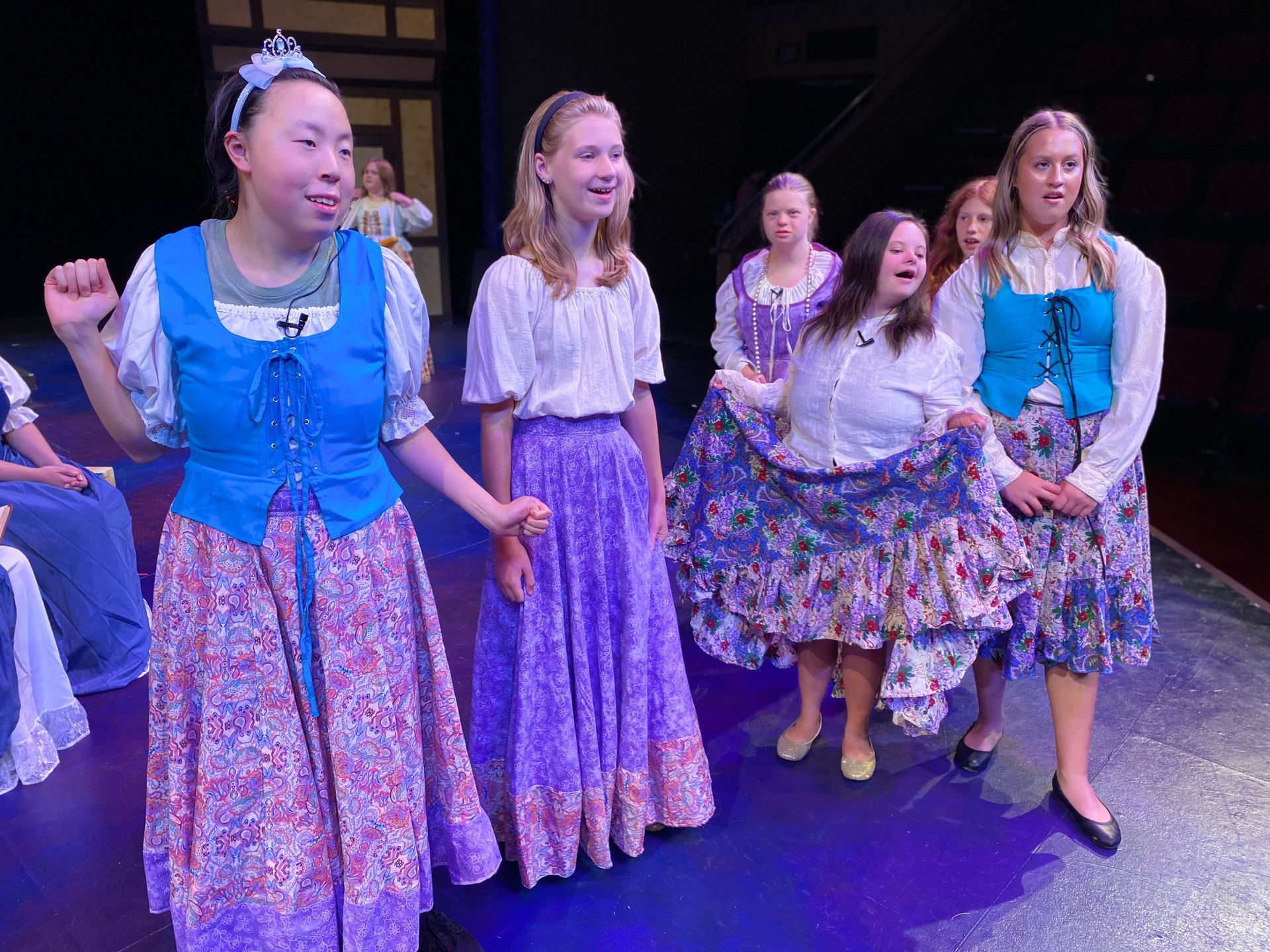Cast members in costume on a stage; one girl in blue top and floral skirt dances.