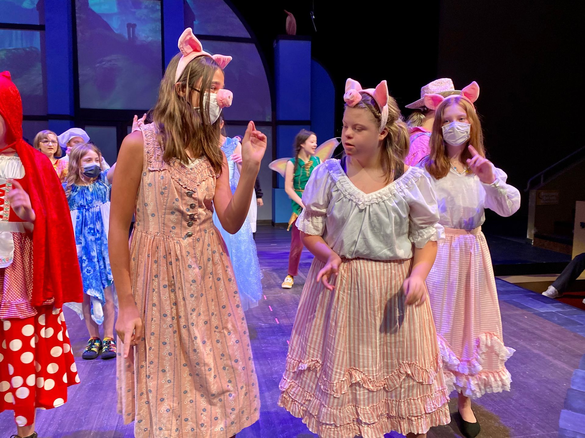 Children in a play, some wearing pig ears. Stage setting, girls talking and gesturing.