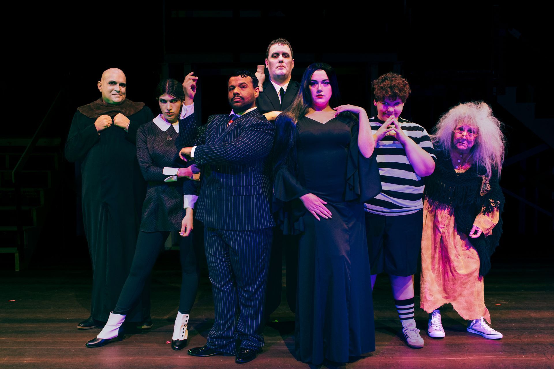 Seven performers in dark stage costumes pose under colorful lighting on a theater stage.