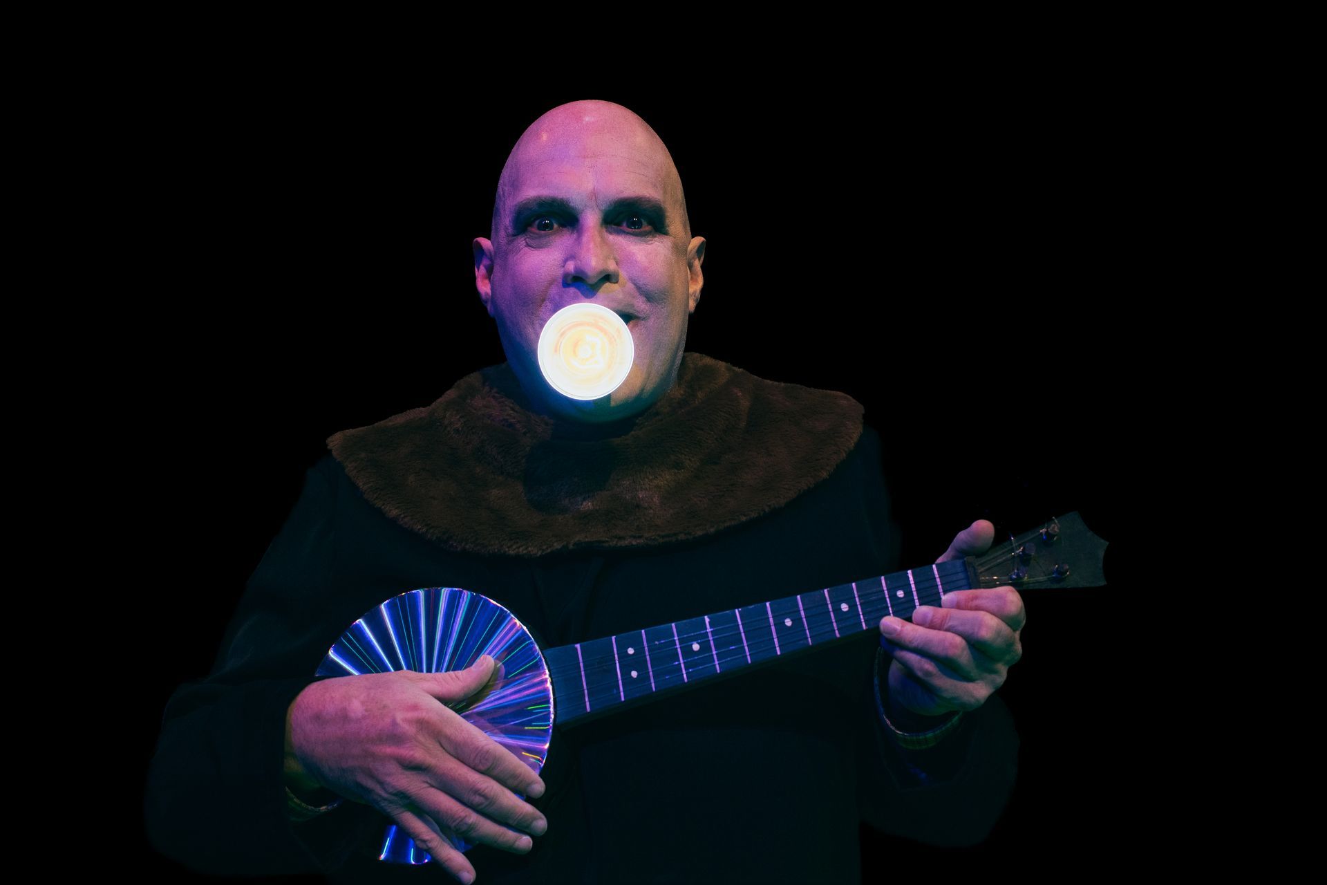 Person in dark clothing playing a blue string instrument with a glowing orb at the mouth against a black background