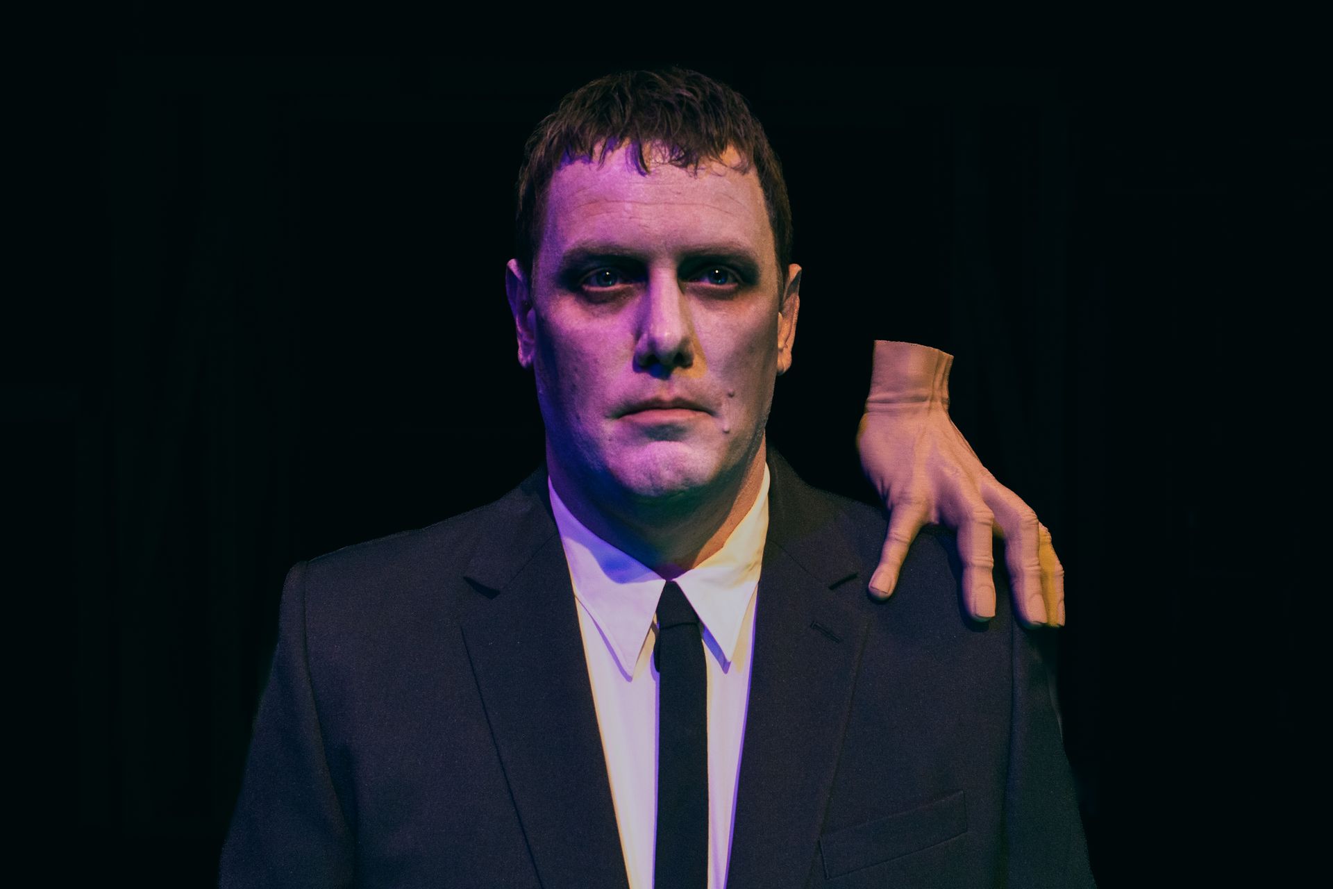 A man in a dark suit stands under purple lighting with a hand resting on his shoulder.