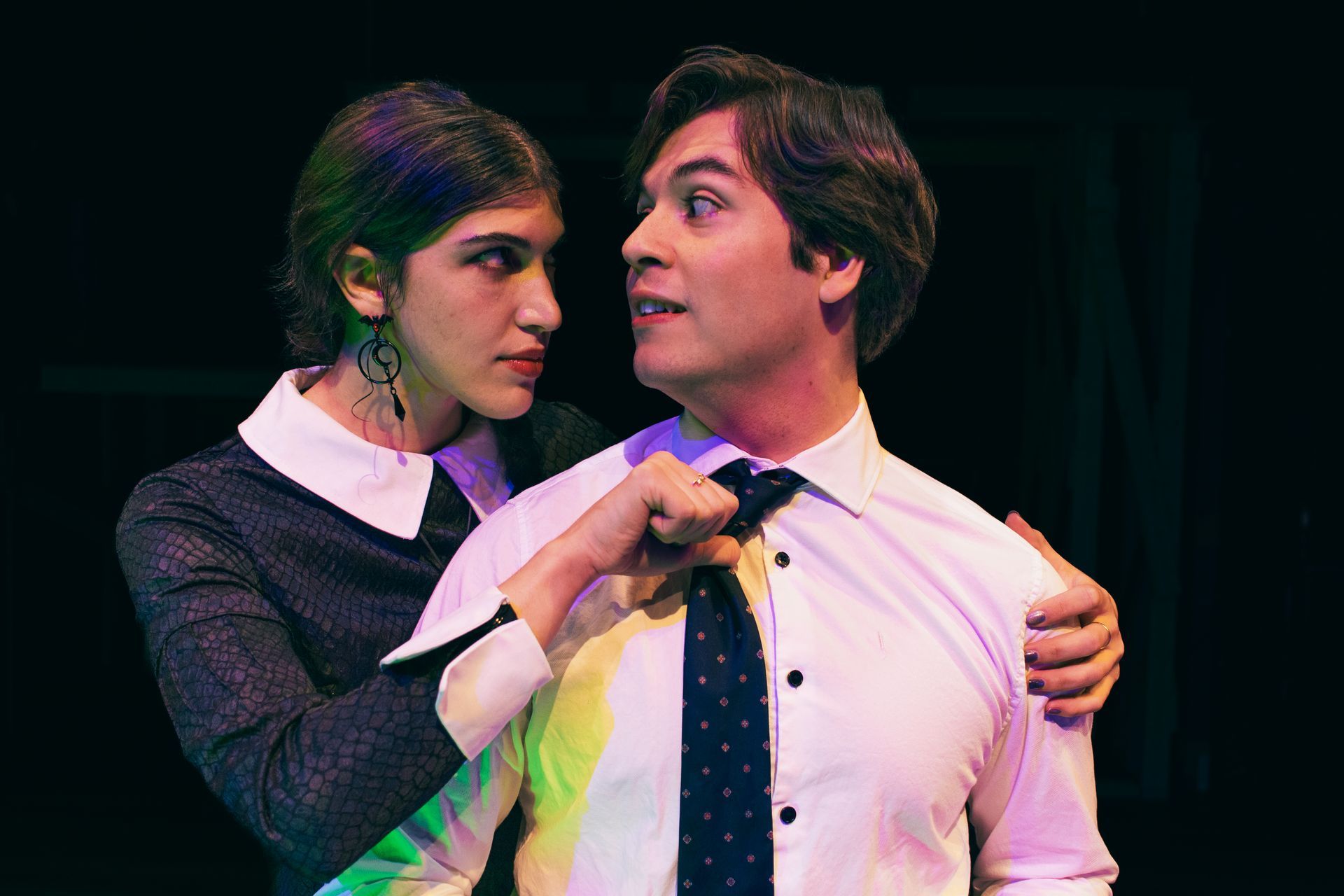 Two people on stage, one adjusting the other’s tie, both looking at each other under colored lighting
