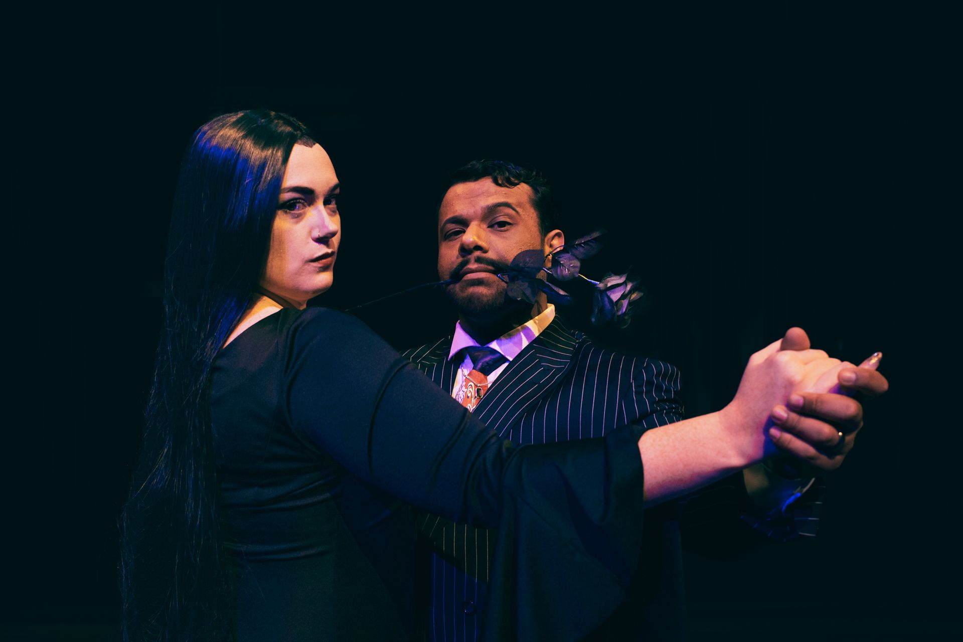Two people in dark formalwear pose together against a dark background, one extending an arm forward.