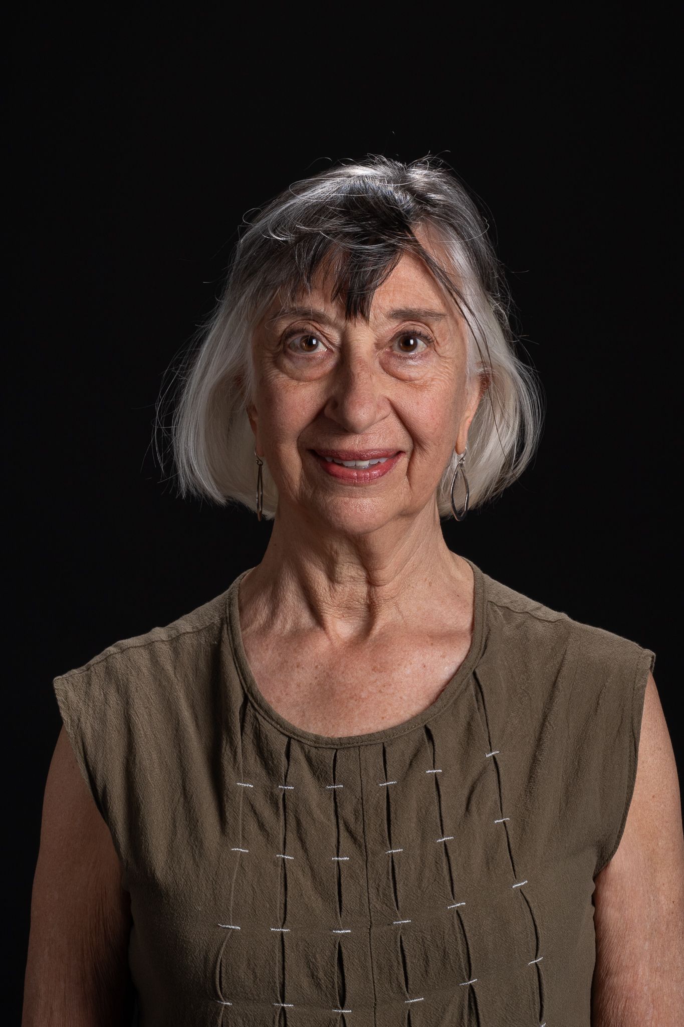 Portrait of a smiling woman with short gray hair against a black background, wearing a brown top