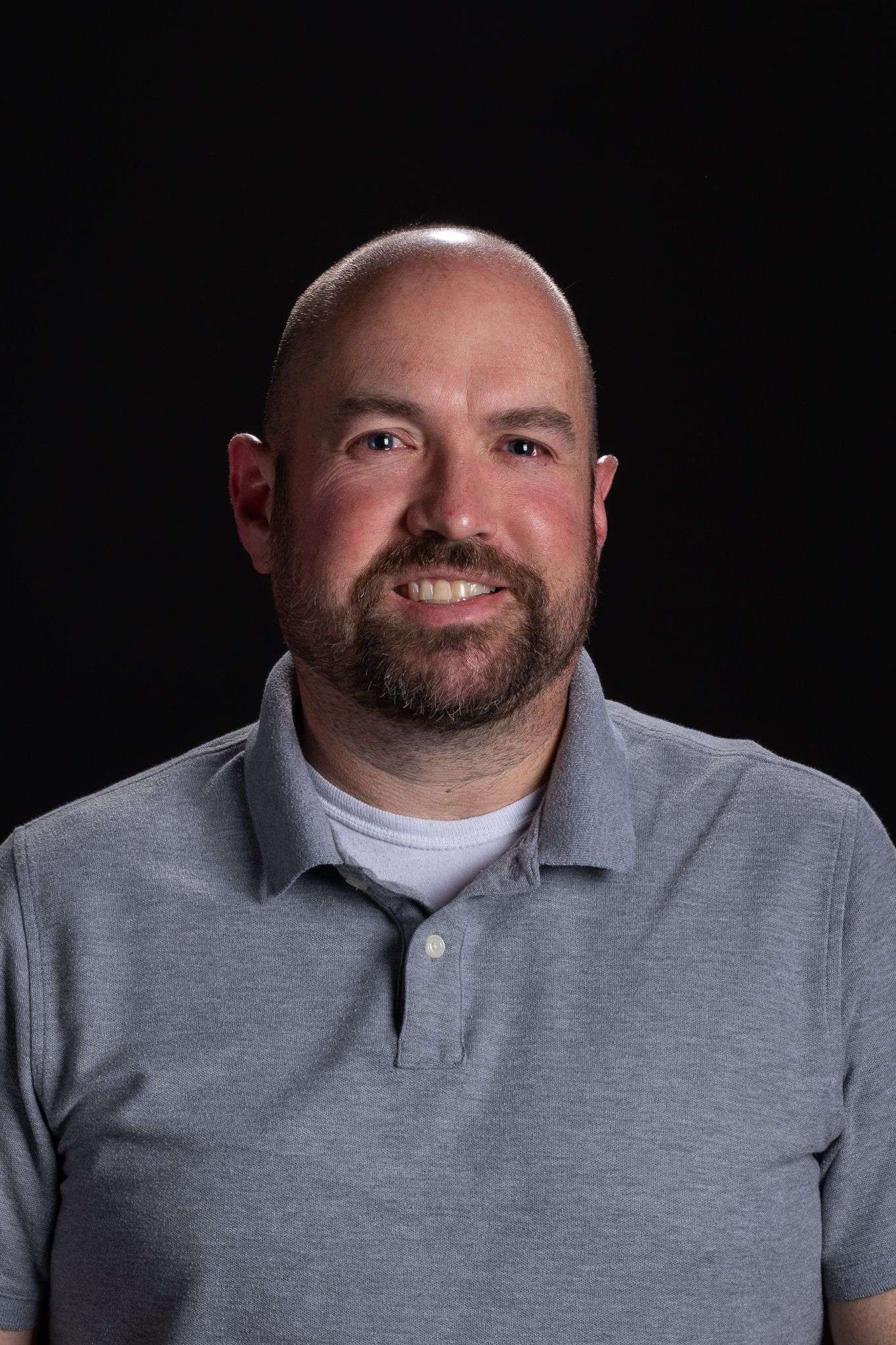 Man in a gray polo shirt against a dark background, smiling at the camera.