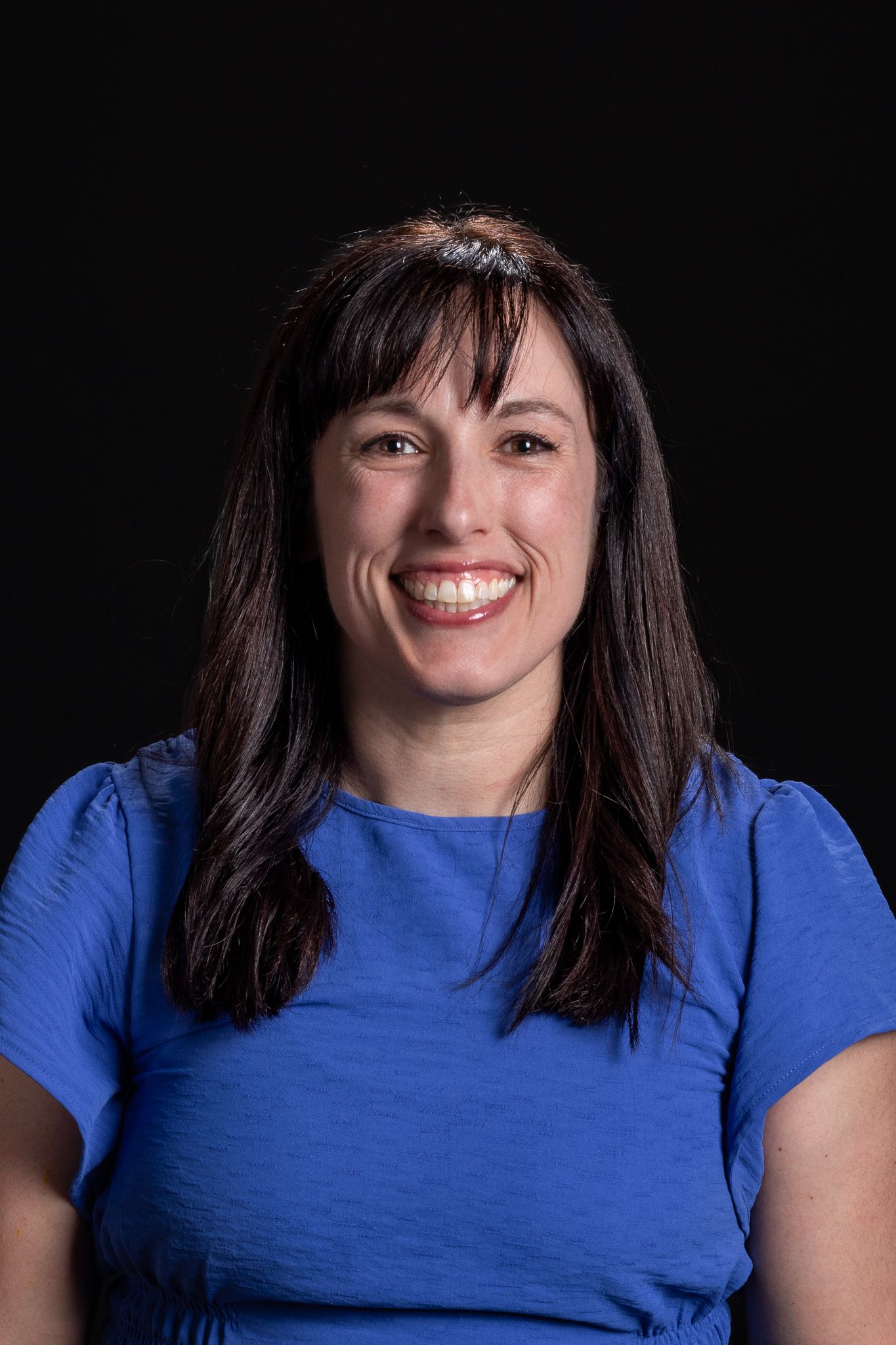 Smiling woman with long dark hair wearing a blue shirt against a black background