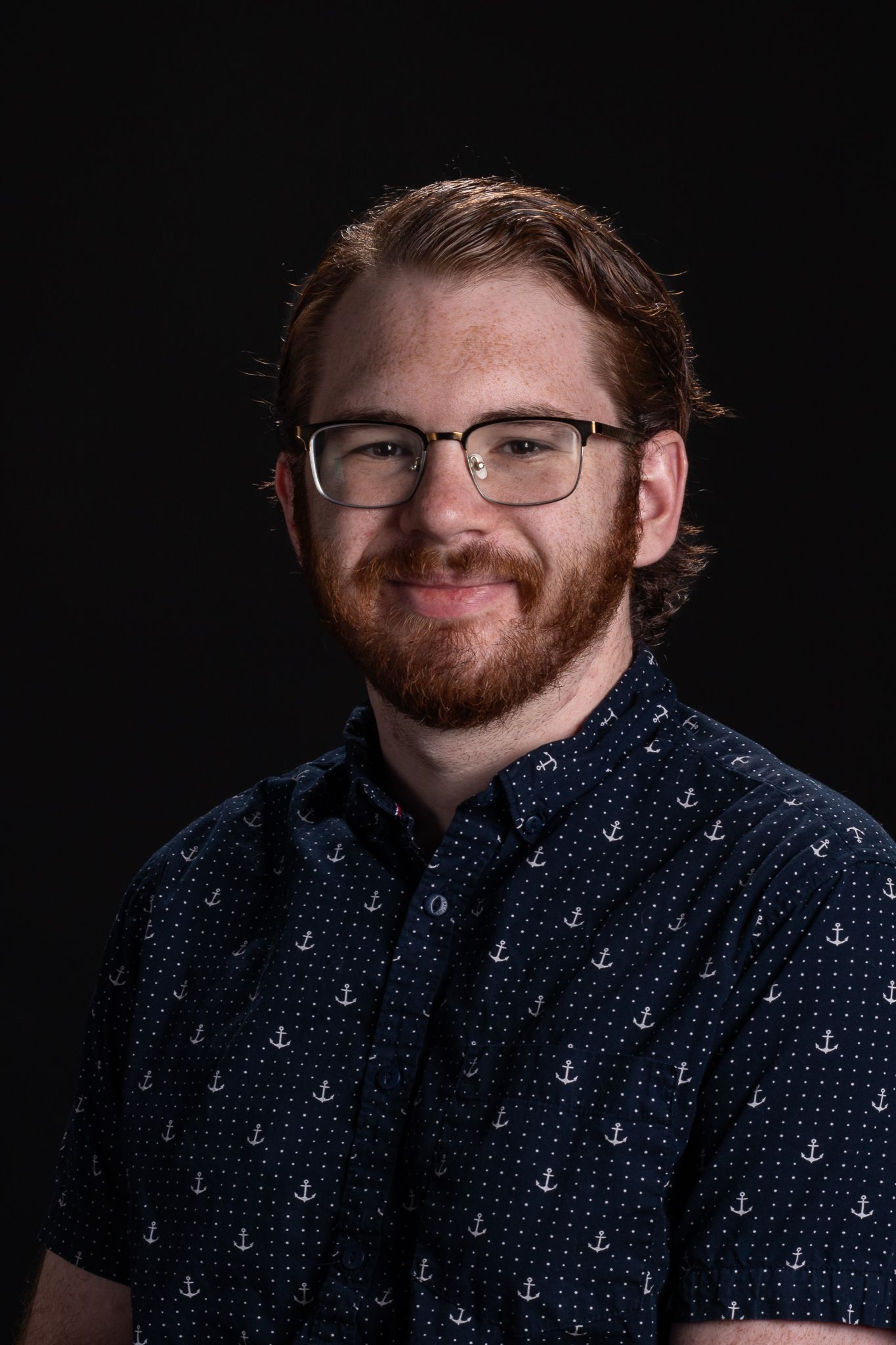 Portrait of a smiling person with glasses, beard, and dark patterned shirt against a black background