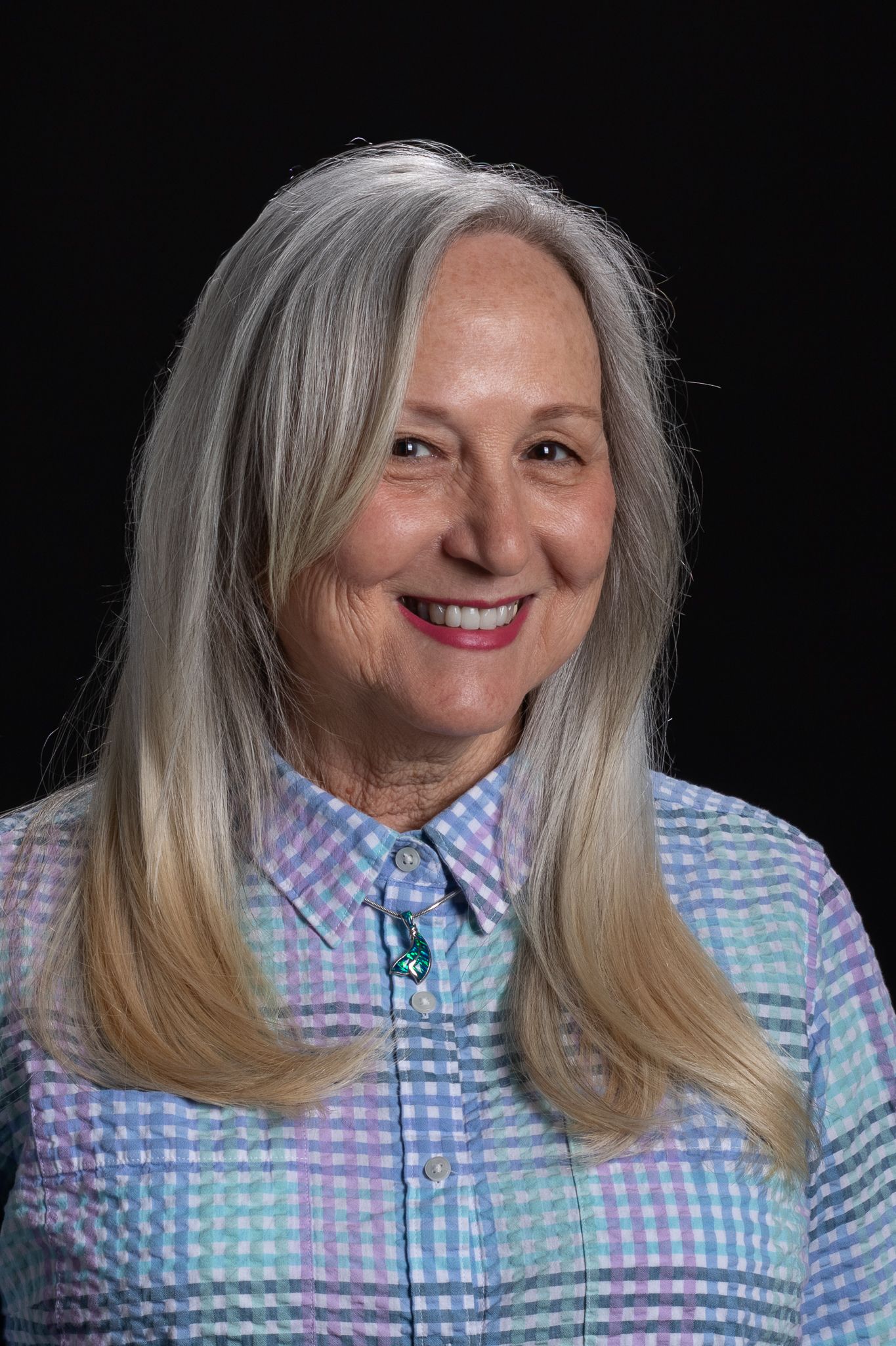 Smiling woman with long gray hair wearing a pastel plaid shirt against a black background