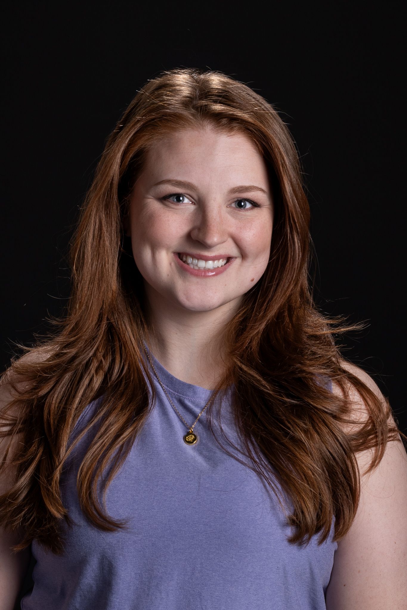 Smiling woman with long brown hair wearing a lavender shirt against a black background