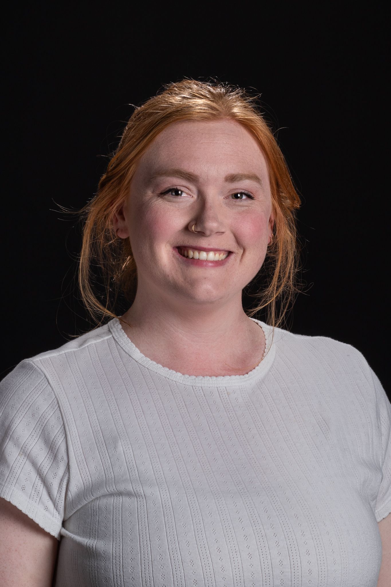 Smiling person with red hair wearing a white shirt against a dark background