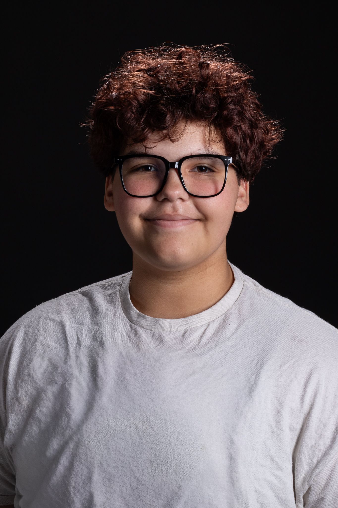 Smiling person with curly hair and glasses, wearing a white shirt against a dark background