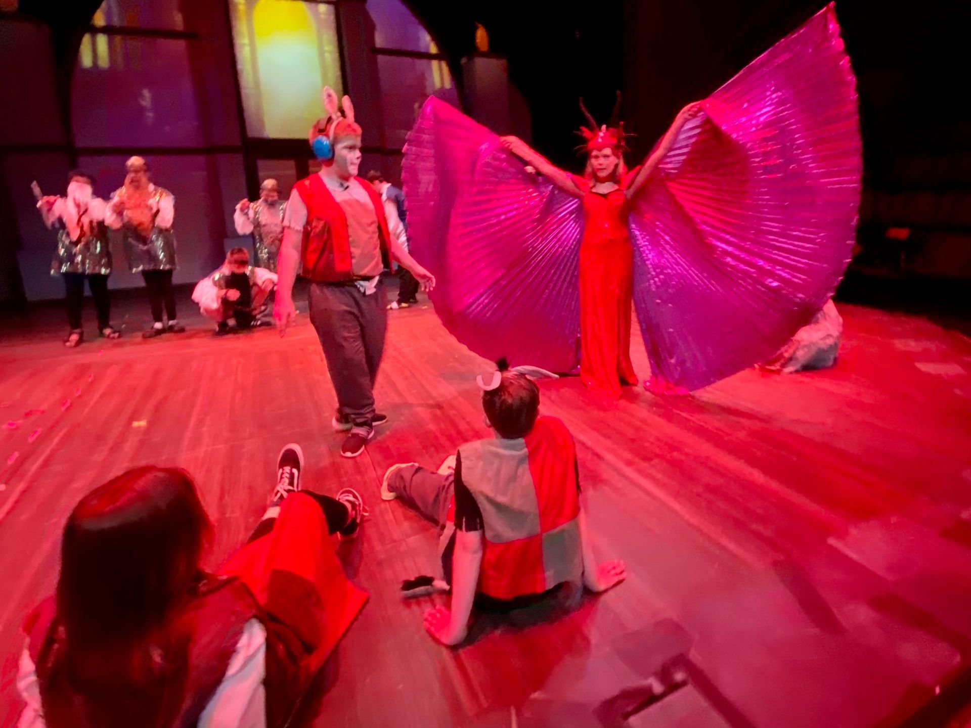 Stage performance; a figure in a red dress with large pink wings; actors in costume on stage with a red lit floor.