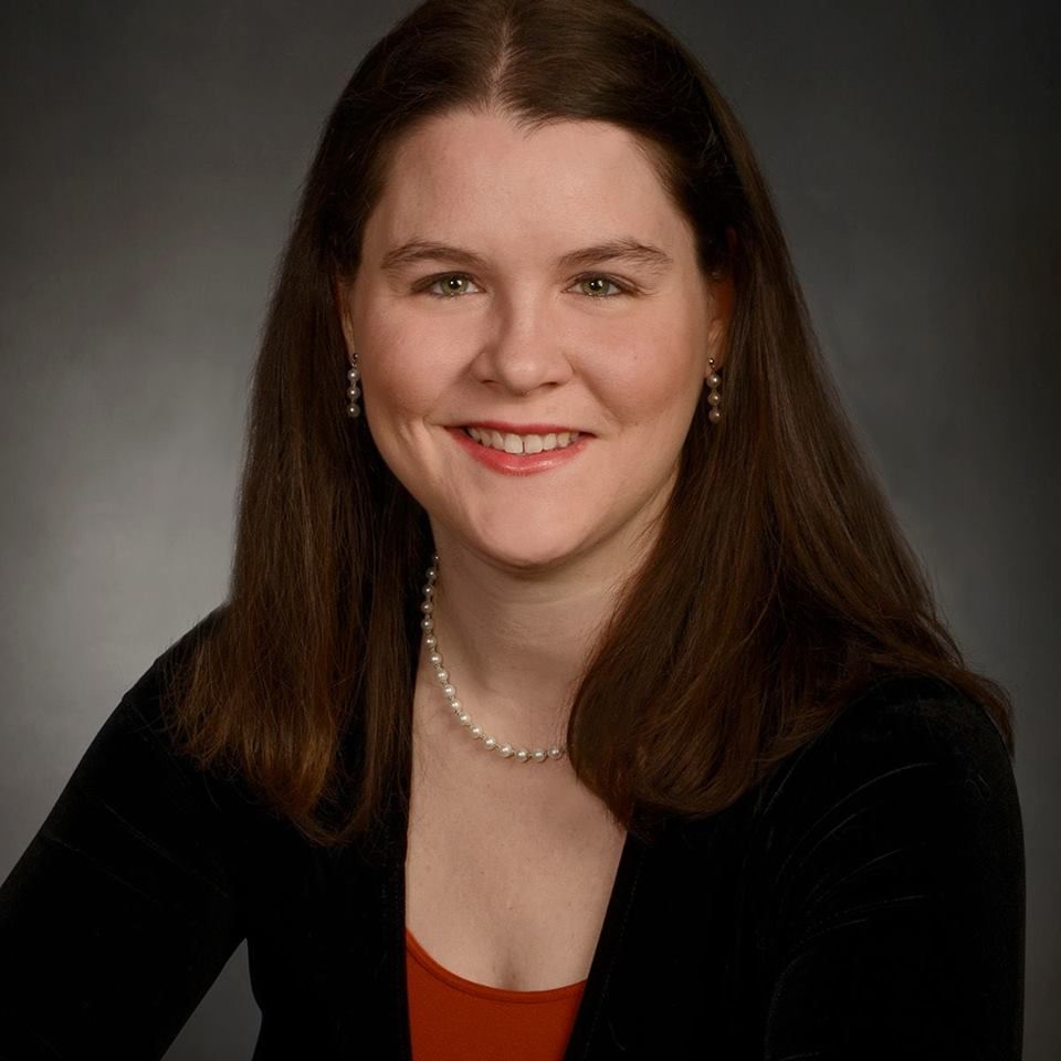 Studio portrait of a smiling woman with long brown hair, wearing a black top and pearl necklace.