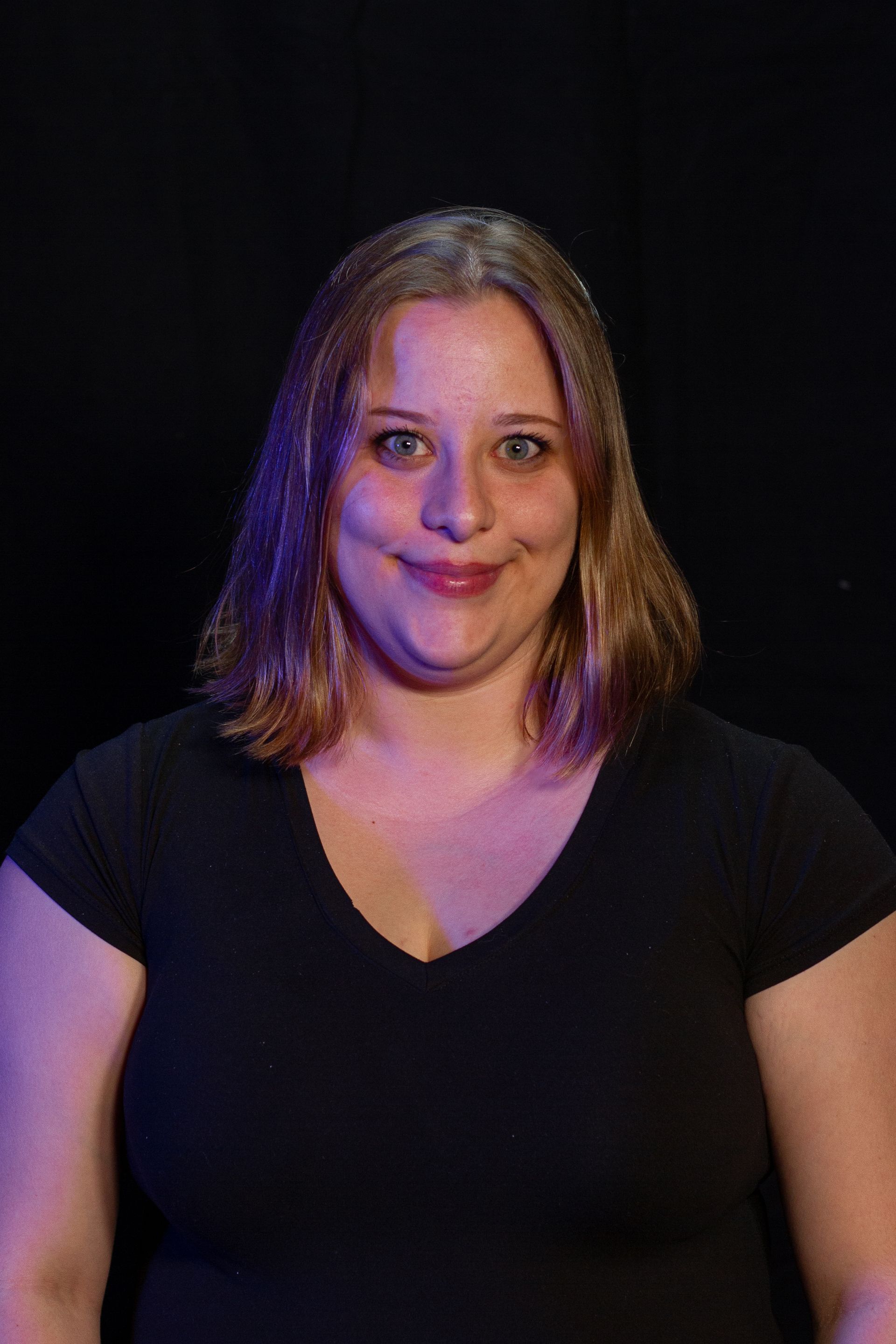 Woman with light brown hair, black shirt, smiles, against a dark background.