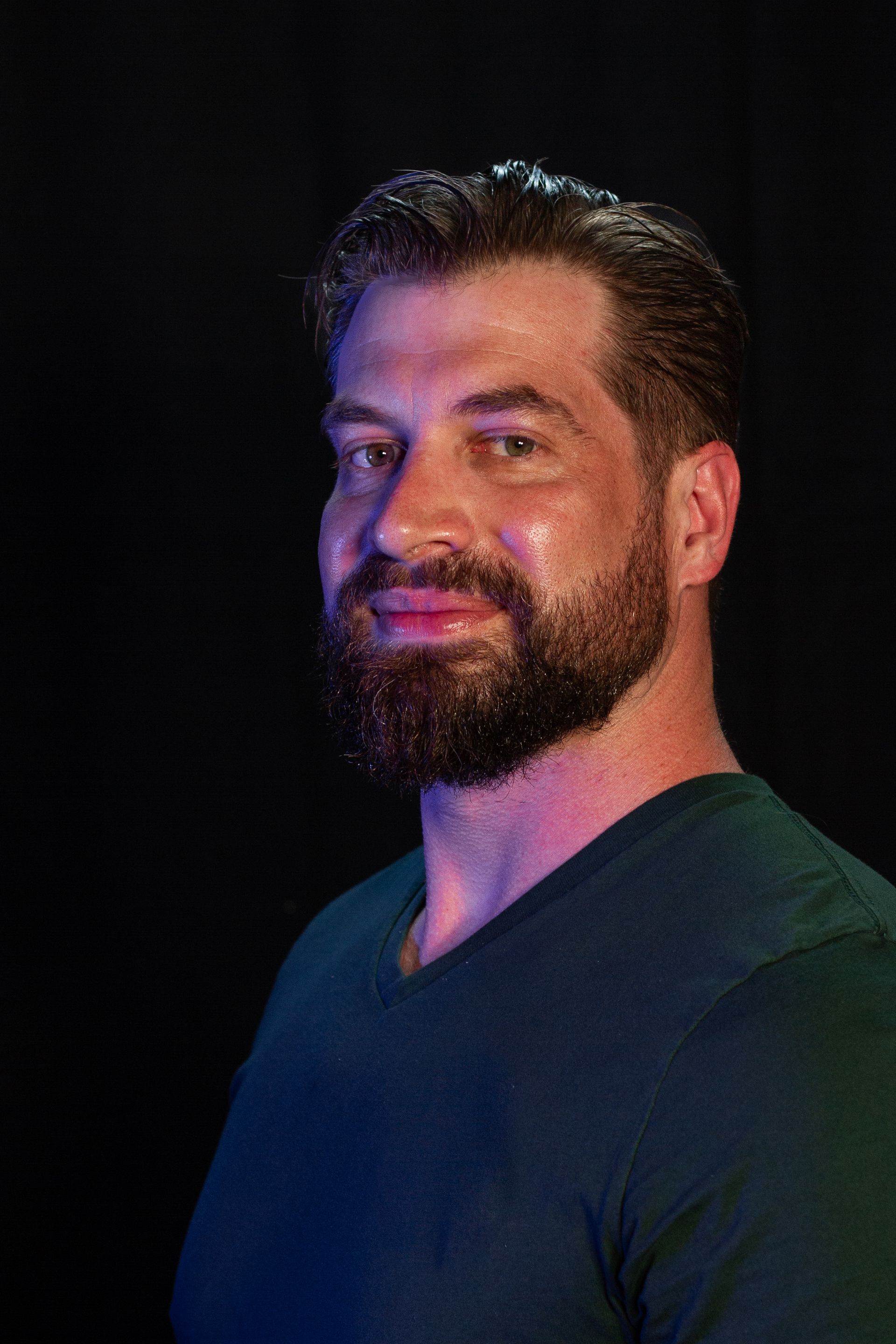 Man with a beard in a dark teal shirt smiles, lit with blue and green light against a black background.
