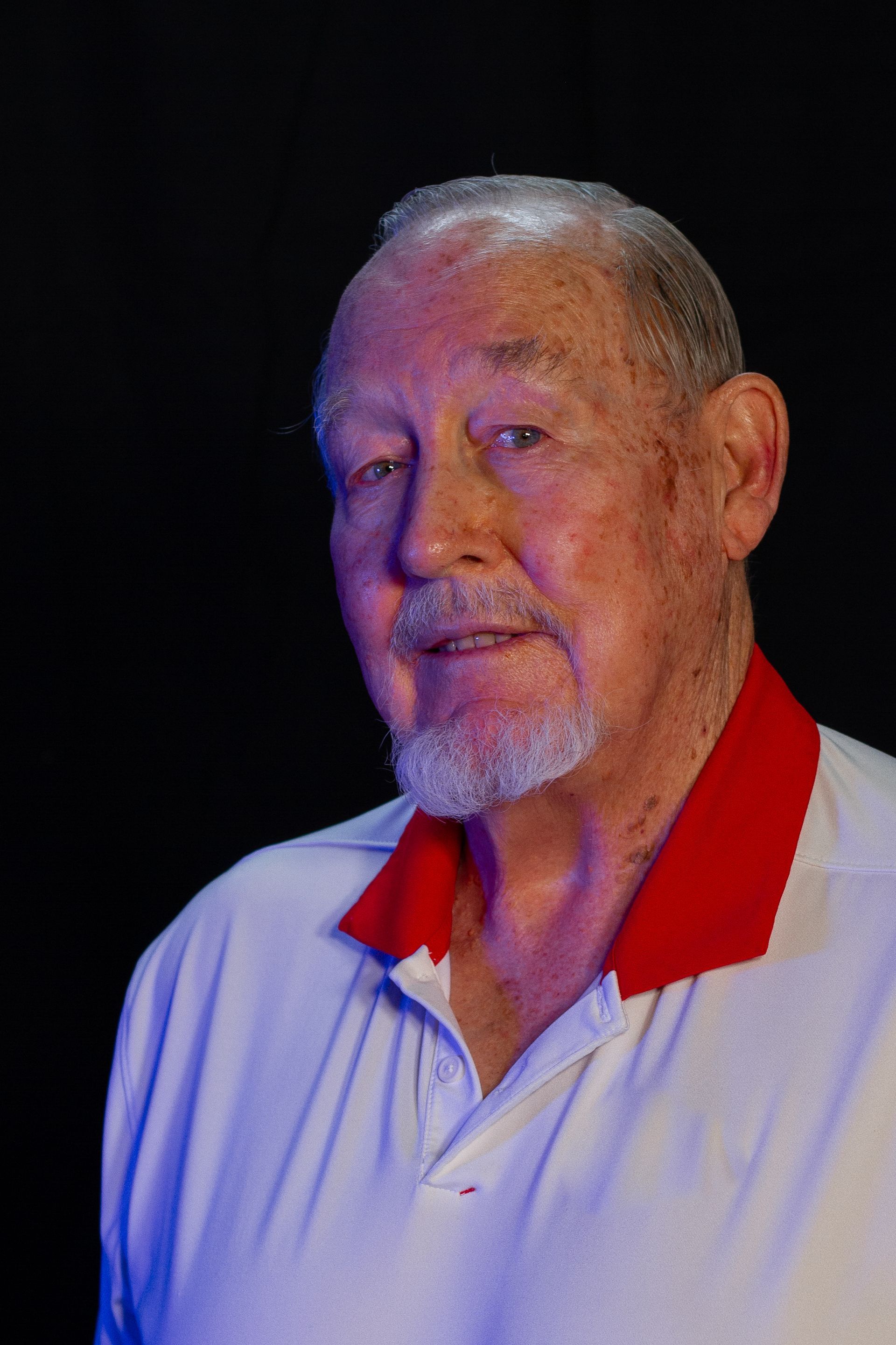 Older man with a white polo shirt with a red collar, short beard, and a neutral expression against a dark background.