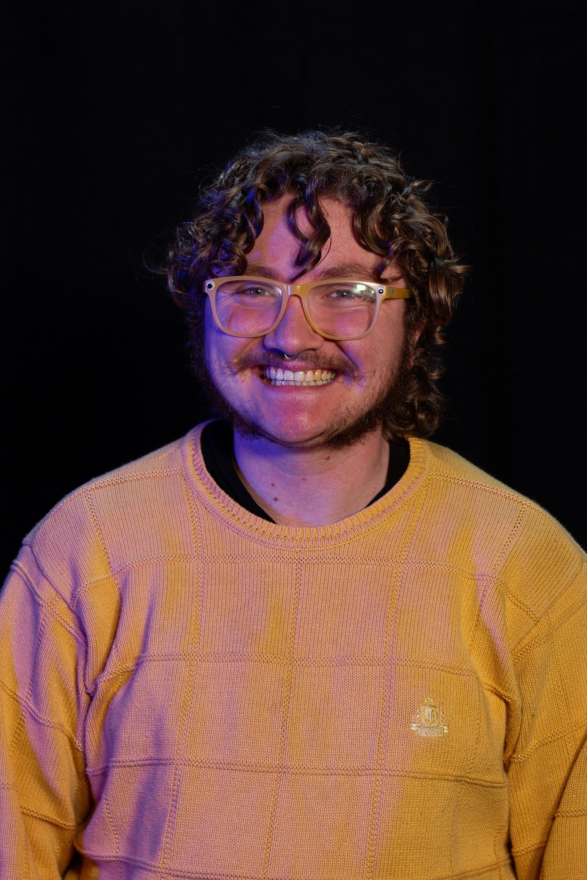 A smiling person with curly brown hair, glasses, and a yellow sweater against a black background.
