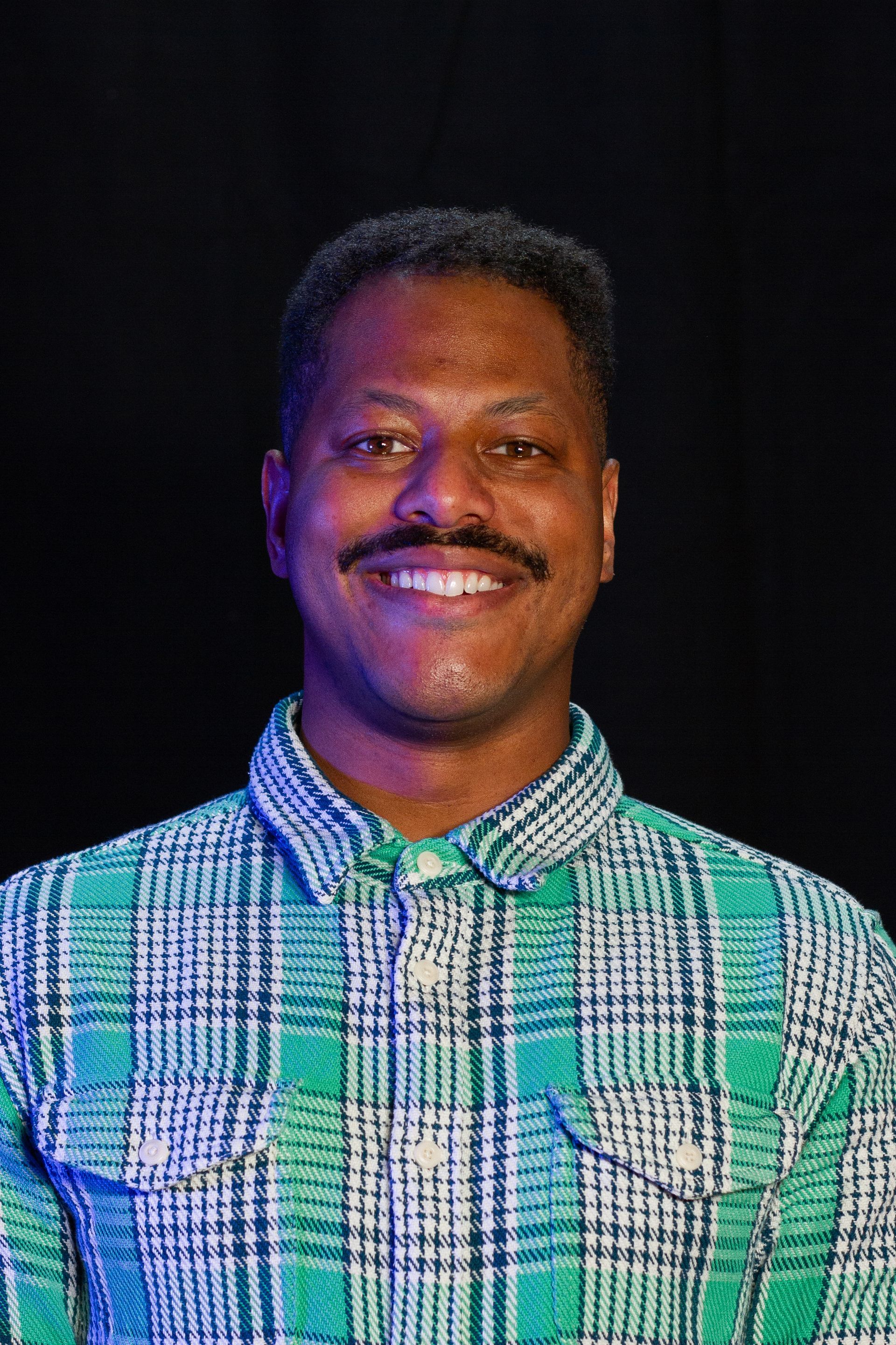 Man with a mustache smiles, wearing a green plaid shirt against a black background.