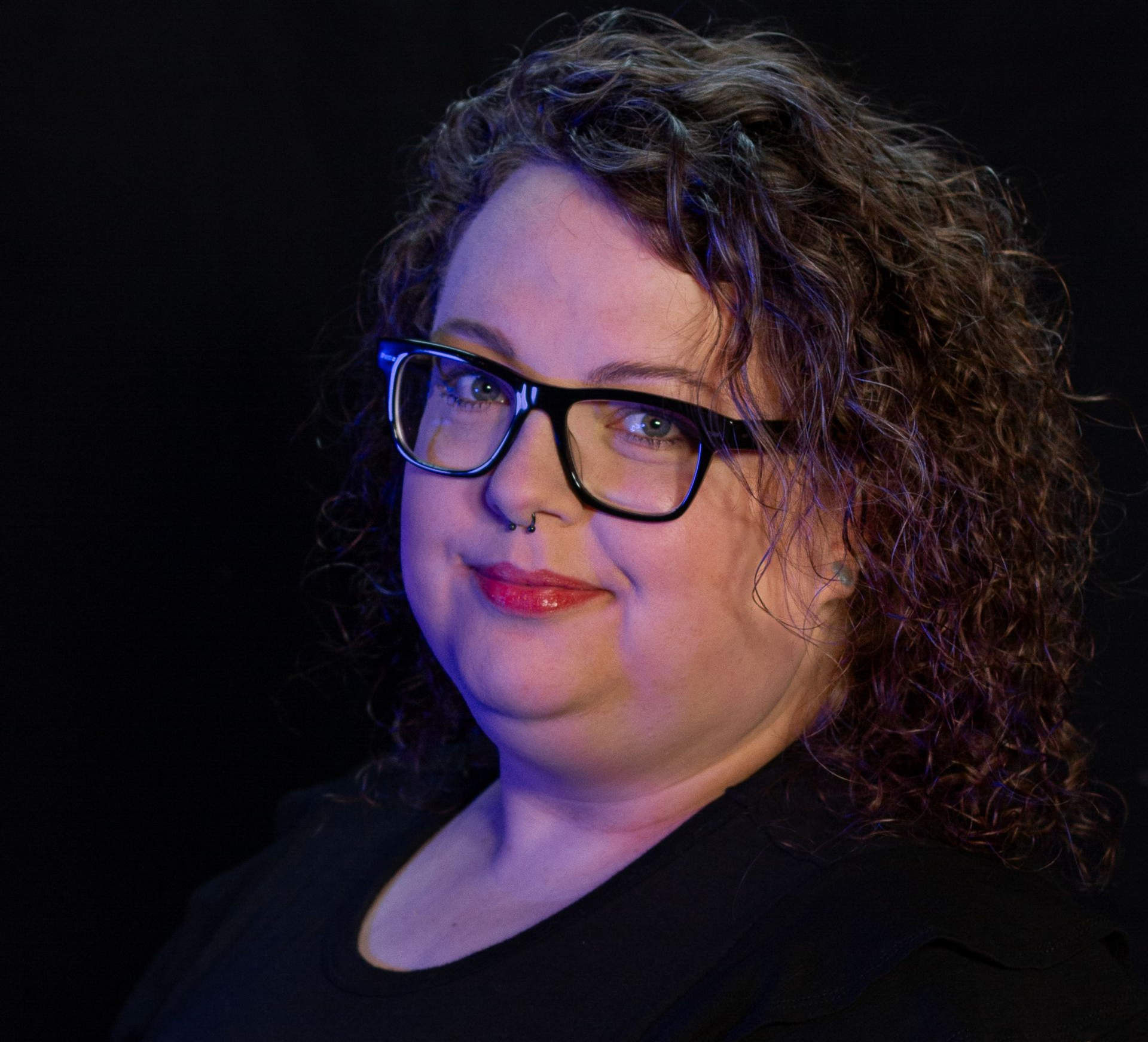 Portrait of a person with curly hair and glasses, softly lit against a dark background