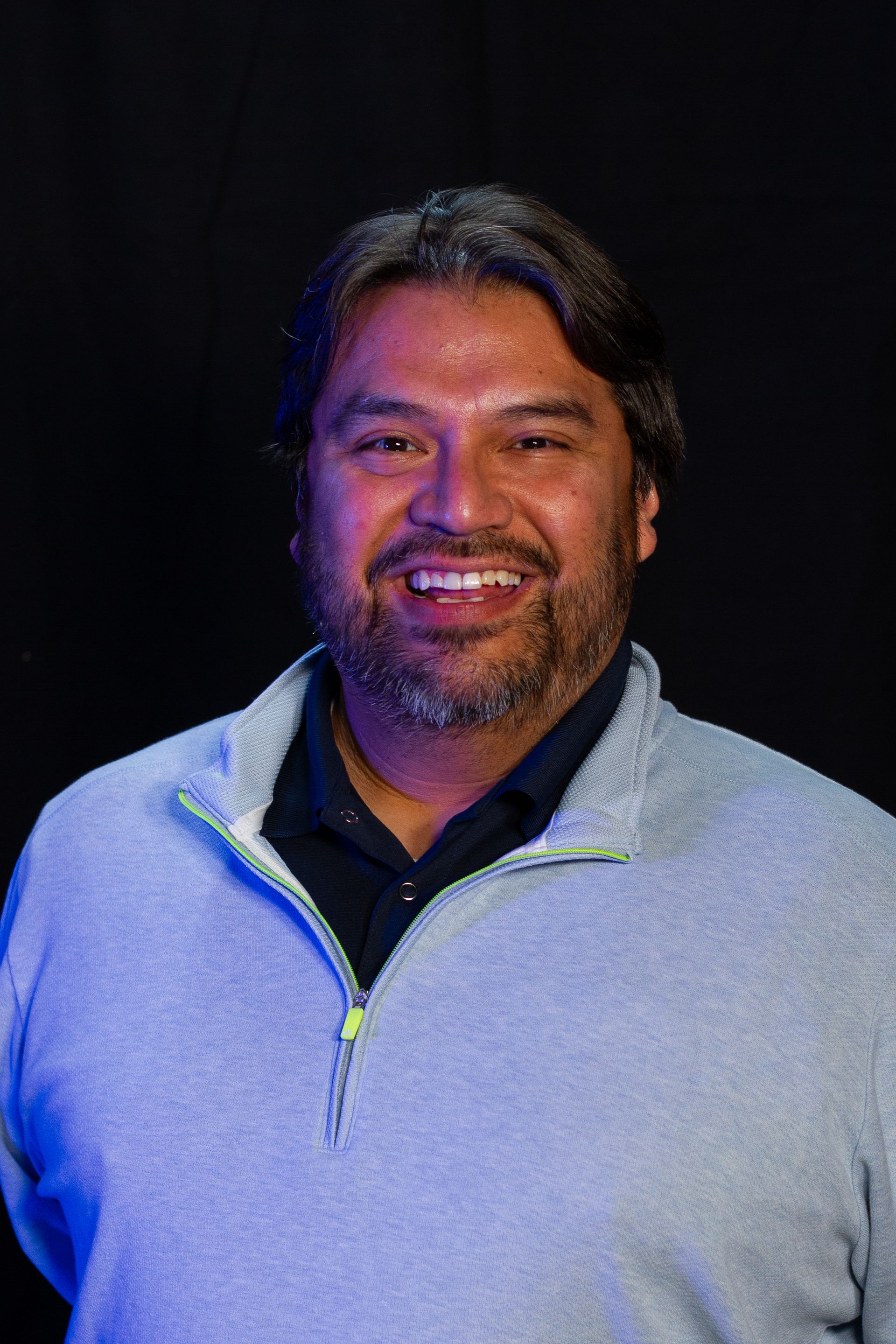 Smiling man with a beard, wearing a light blue sweater, standing against a black backdrop.