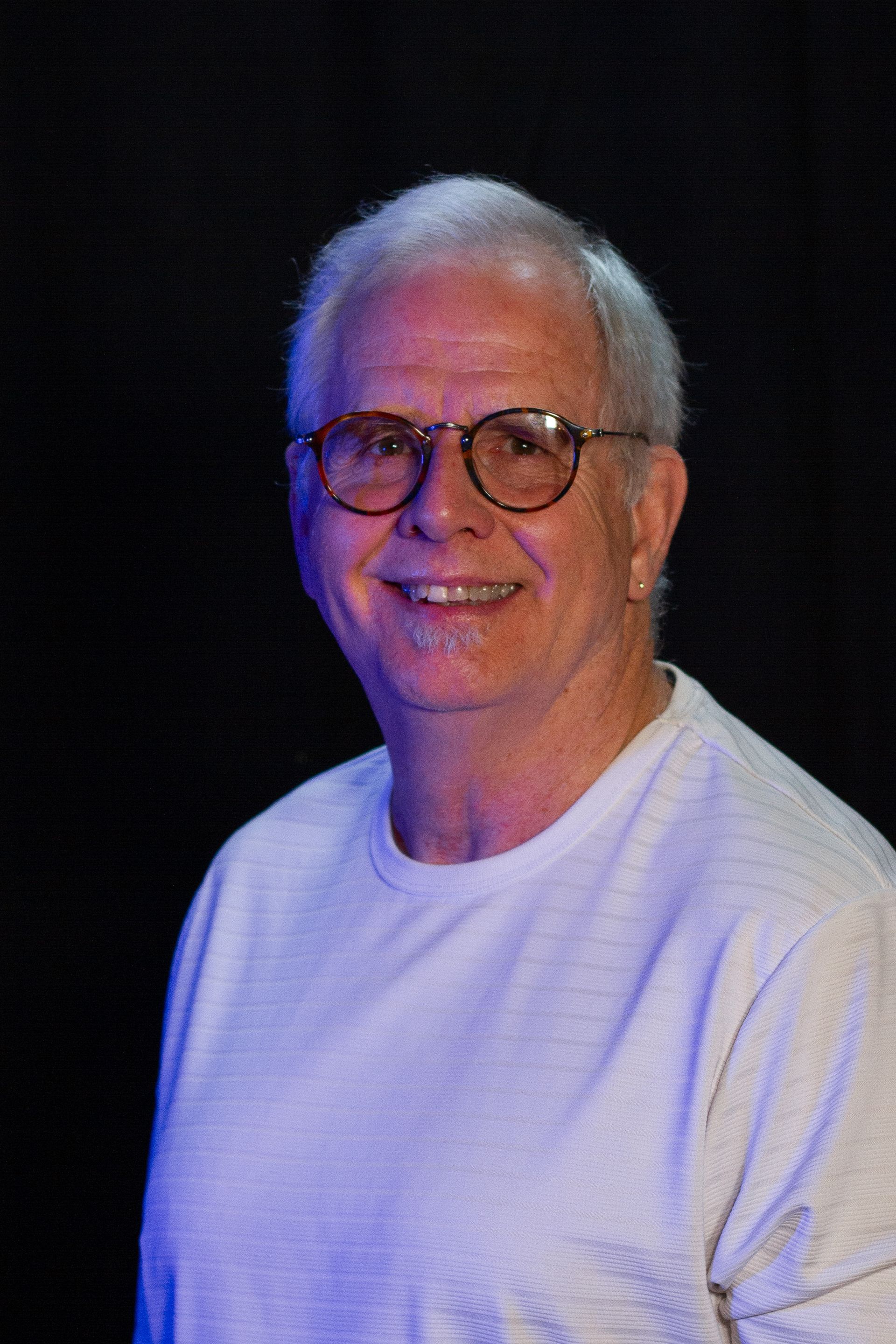 Smiling older man with glasses, white shirt against a black background.