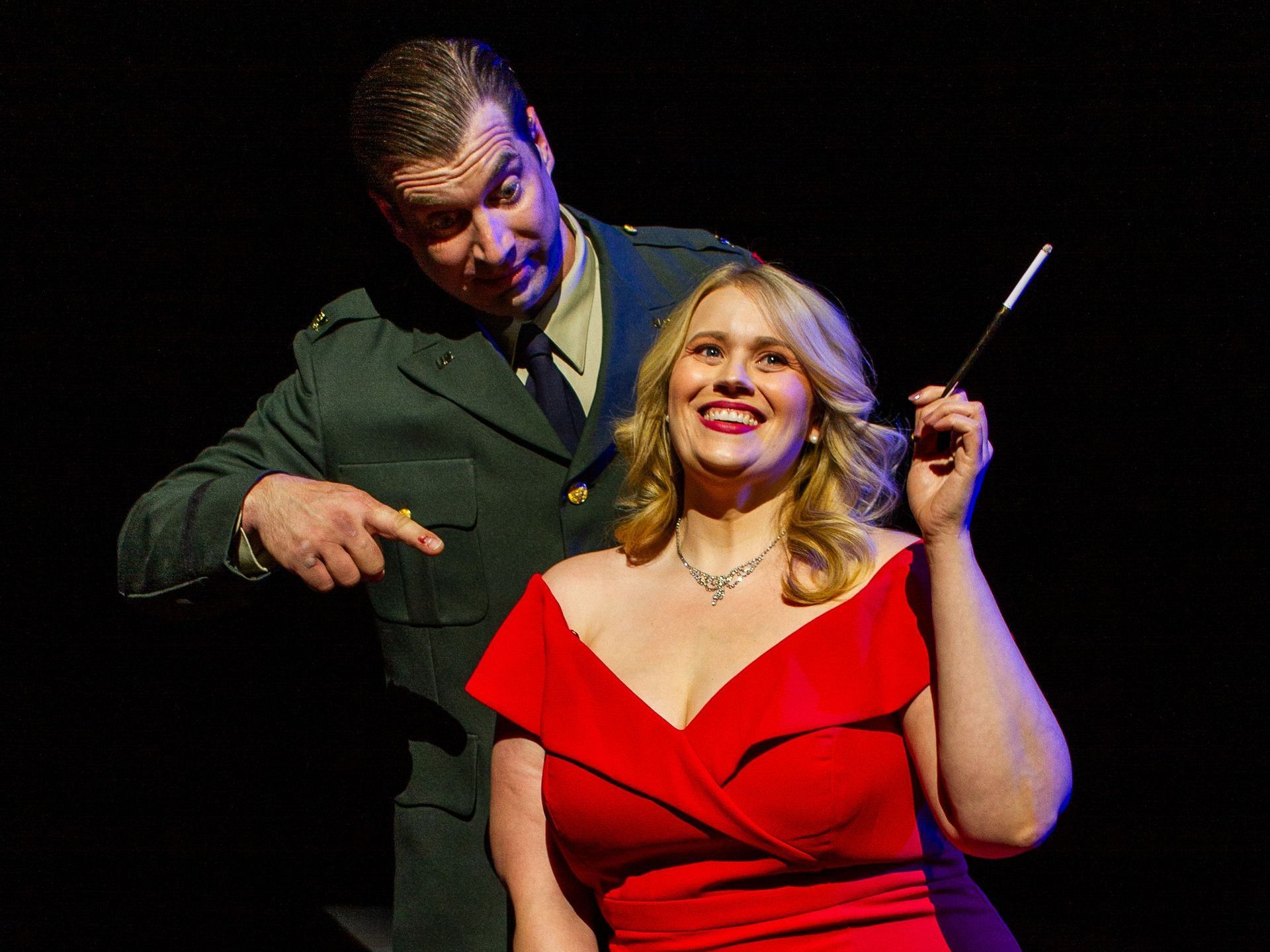 Man in uniform points at woman in red dress, holding cigarette, smiling. Dark background.