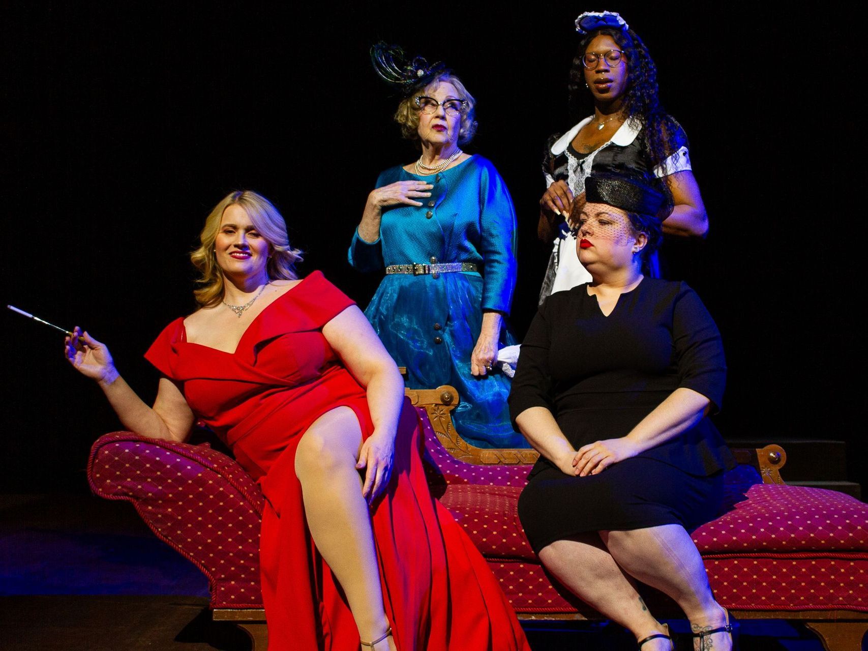 Four women in vintage attire pose on stage. One reclines in red, smoking. Others wear blue, black, and a maid's uniform.