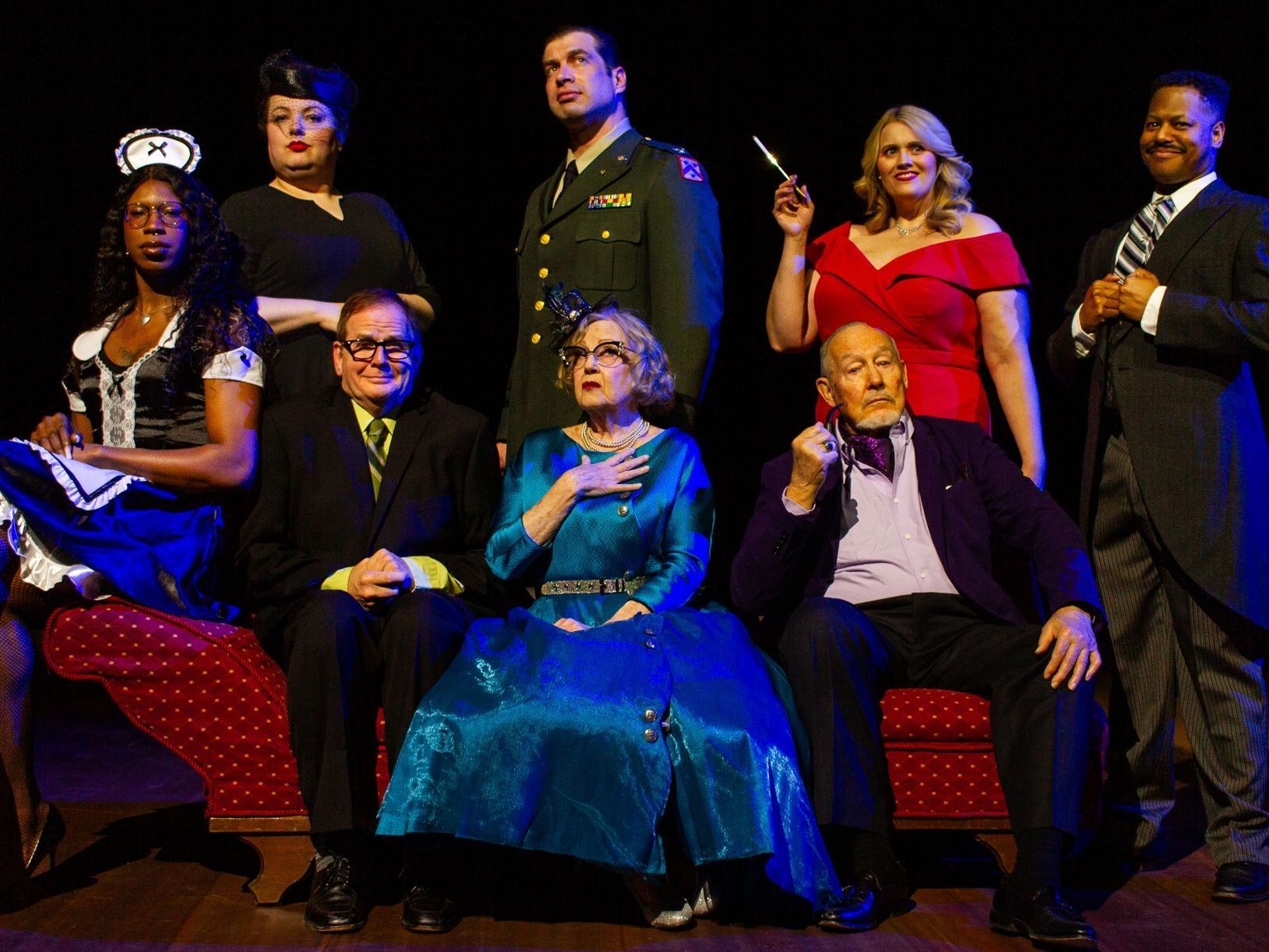 A group of diverse actors in costume on a stage. Some hold props, with varied expressions, lit dramatically.