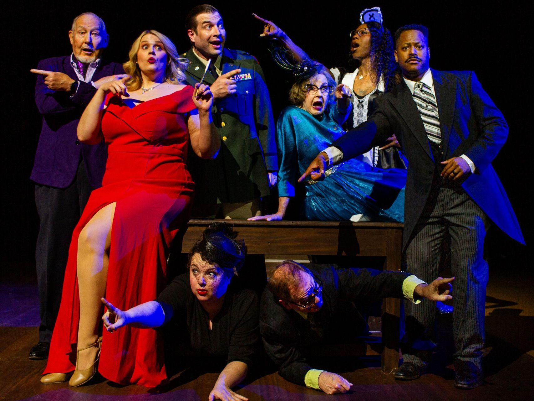 Actors in costume striking poses, some pointing. Varying expressions on faces. Dark stage, studio lighting.