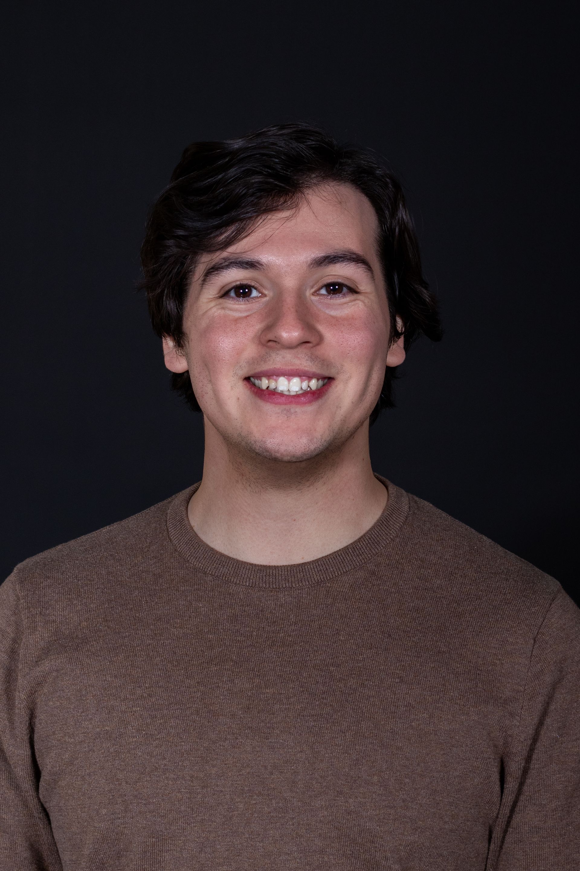 Man with dark hair smiling at the camera, wearing a brown sweater, against a dark gray background.
