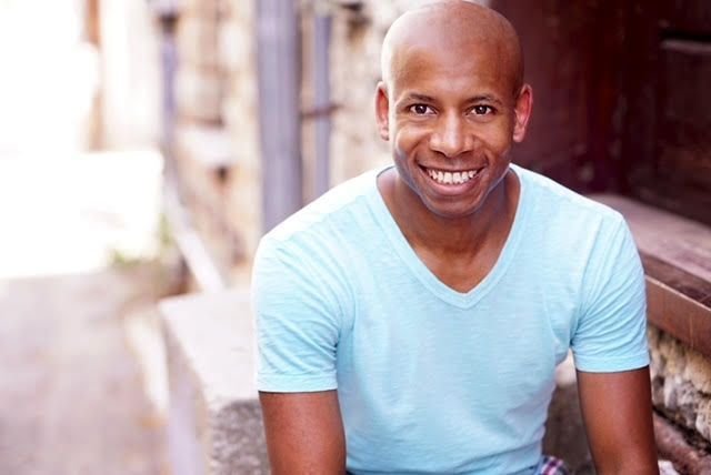 Bald man in a blue v-neck smiling, seated outdoors.