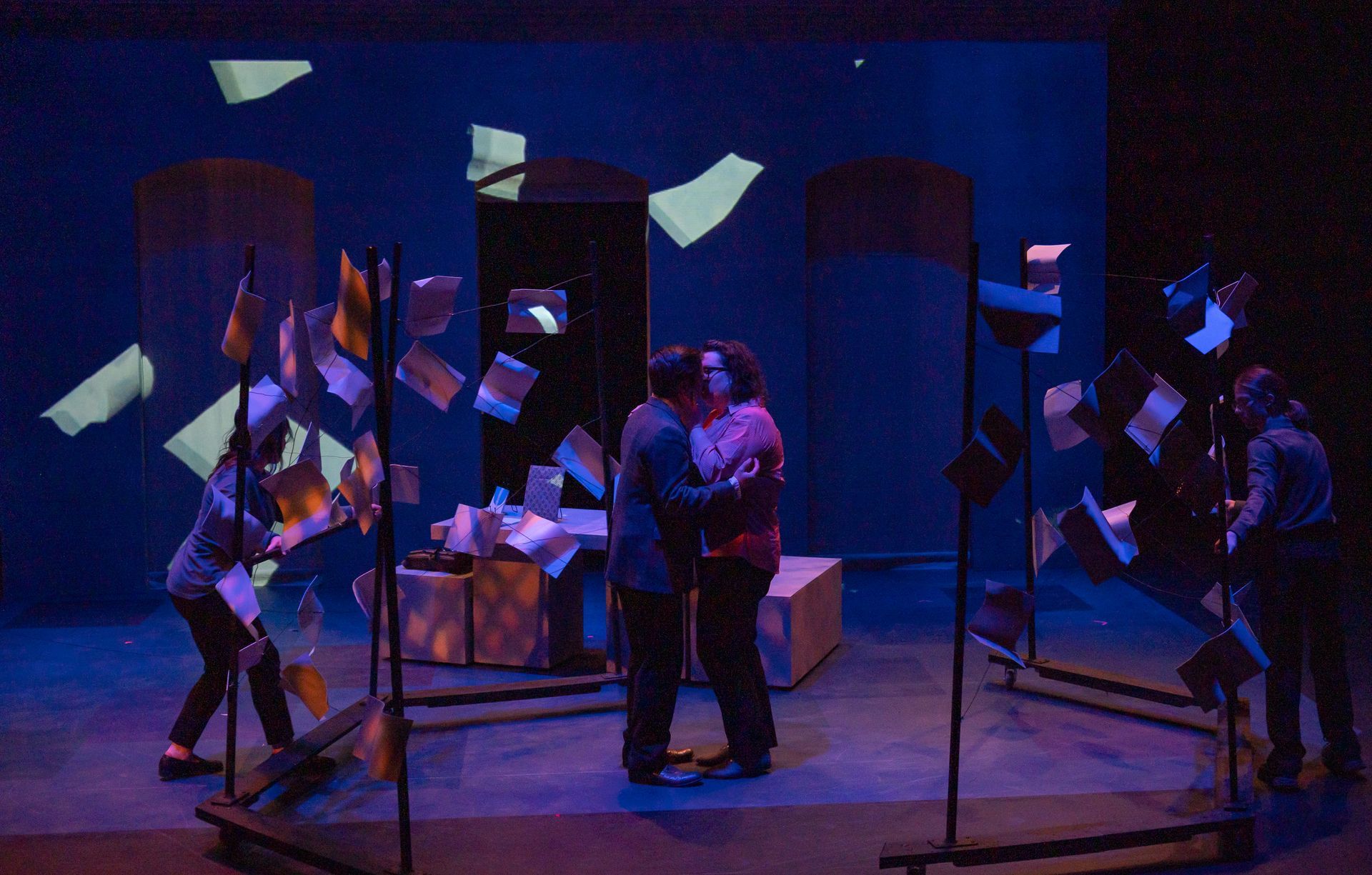 Theater scene: Two embrace center stage surrounded by flag-like props, actors holding flags, blue lighting.