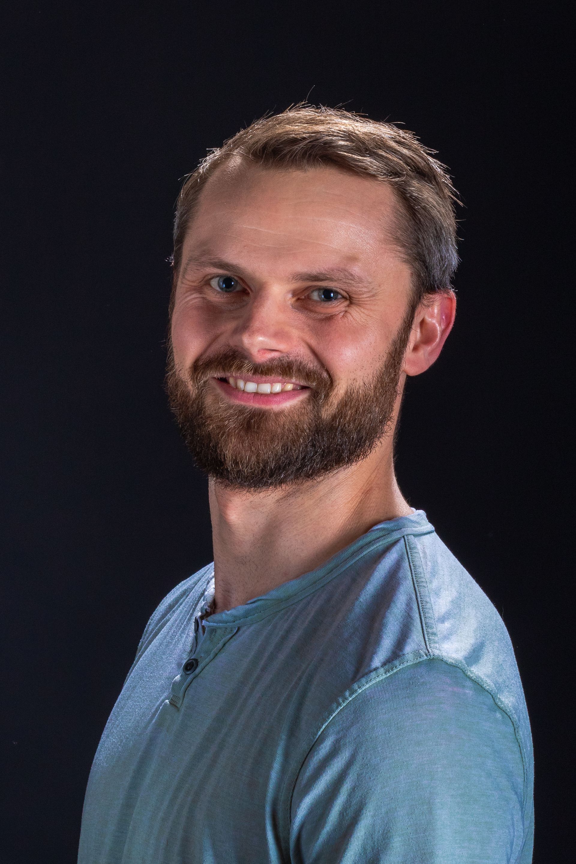 A person with a beard, wearing a blue textured shirt, smiles at the camera against a dark background.
