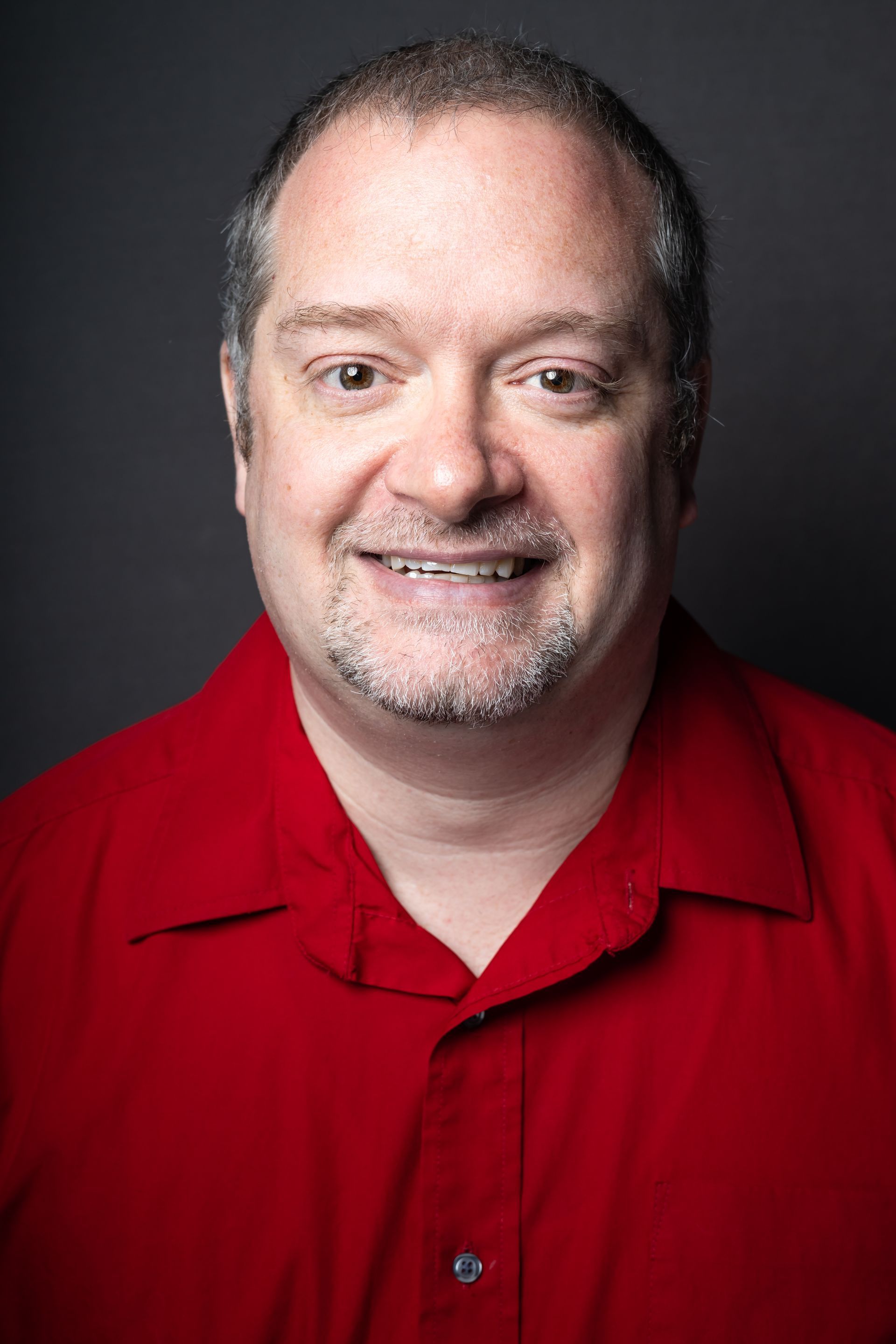 Man with a goatee smiling in a red shirt, against a dark gray background.