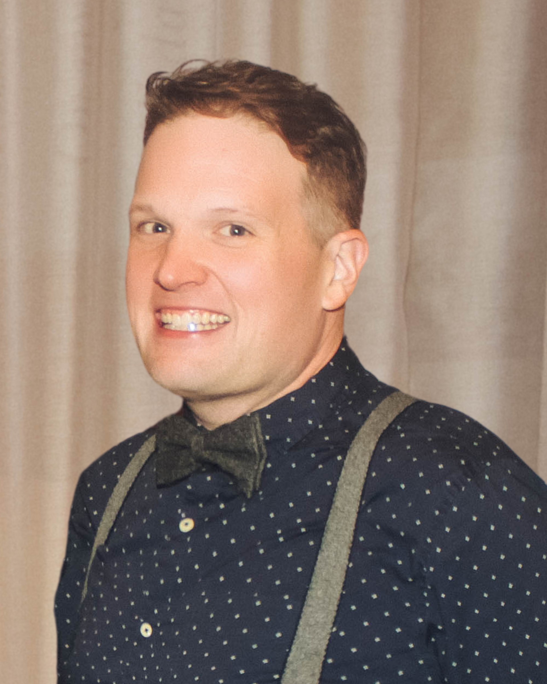 Man smiling, wearing patterned shirt, bow tie, and suspenders.