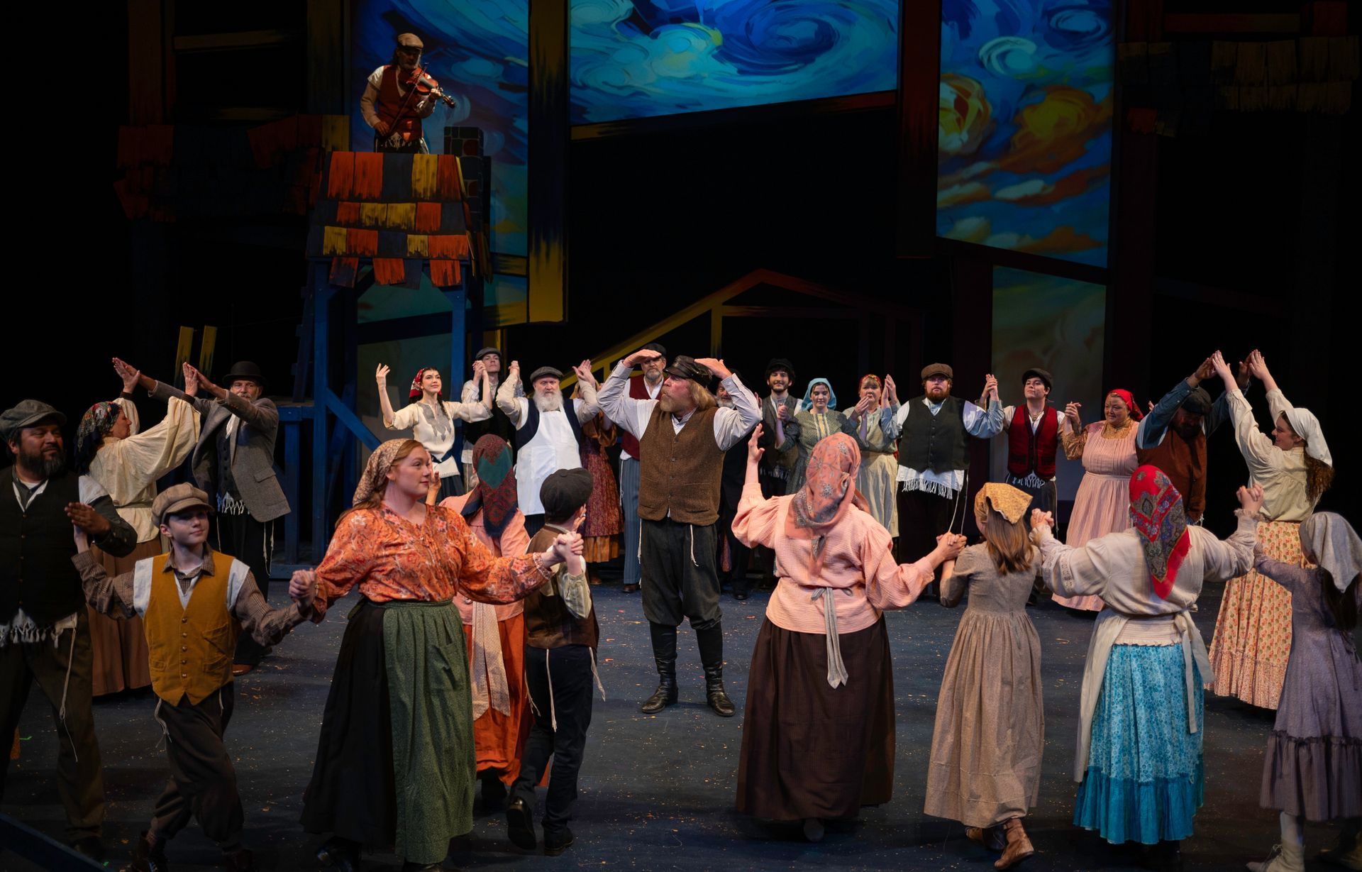 Ensemble cast dances on stage; rustic costumes, hands linked. Colorful backdrop, bright lights.