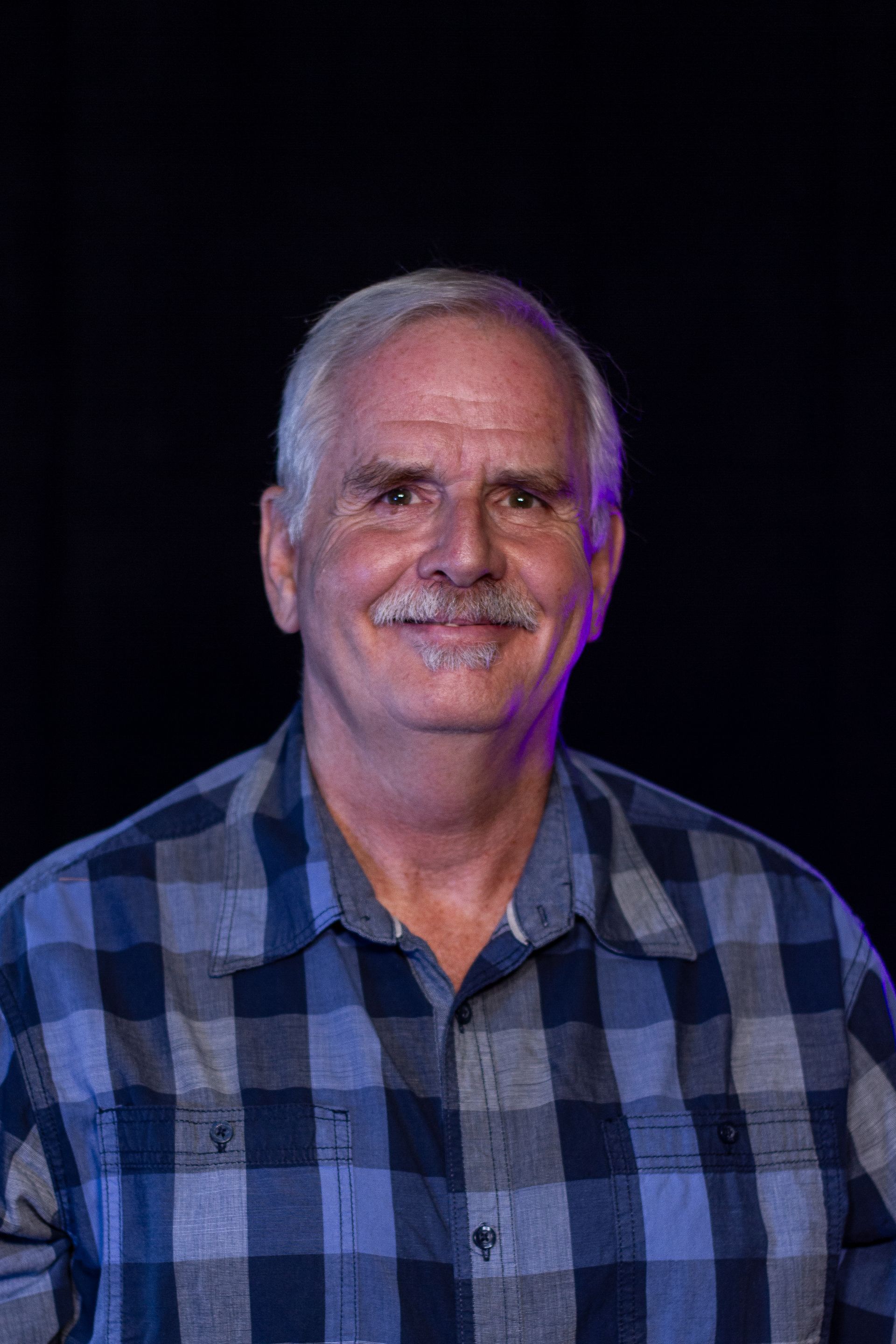 Man with gray hair and mustache smiles in a blue plaid shirt, dark background.