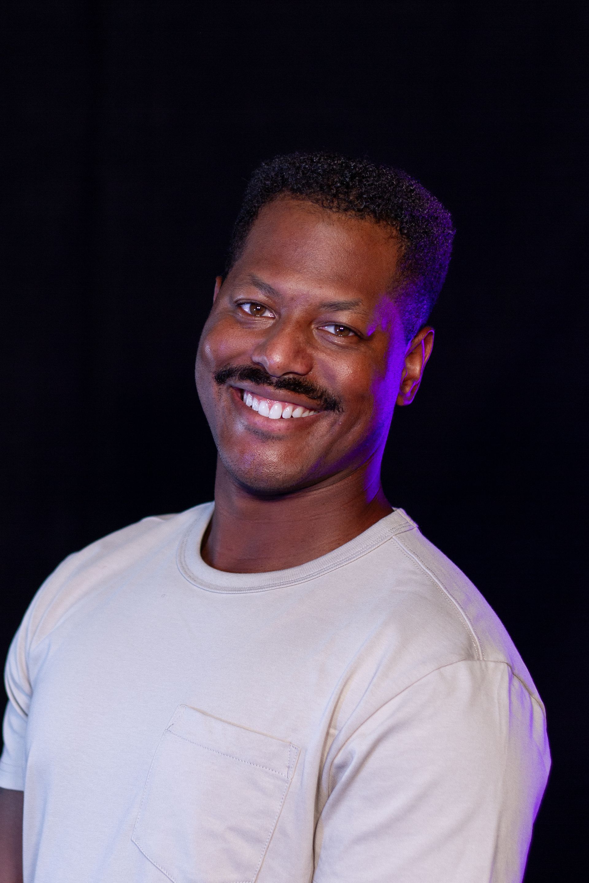 Smiling Black man in a white shirt, set against a black background, with a purple light.