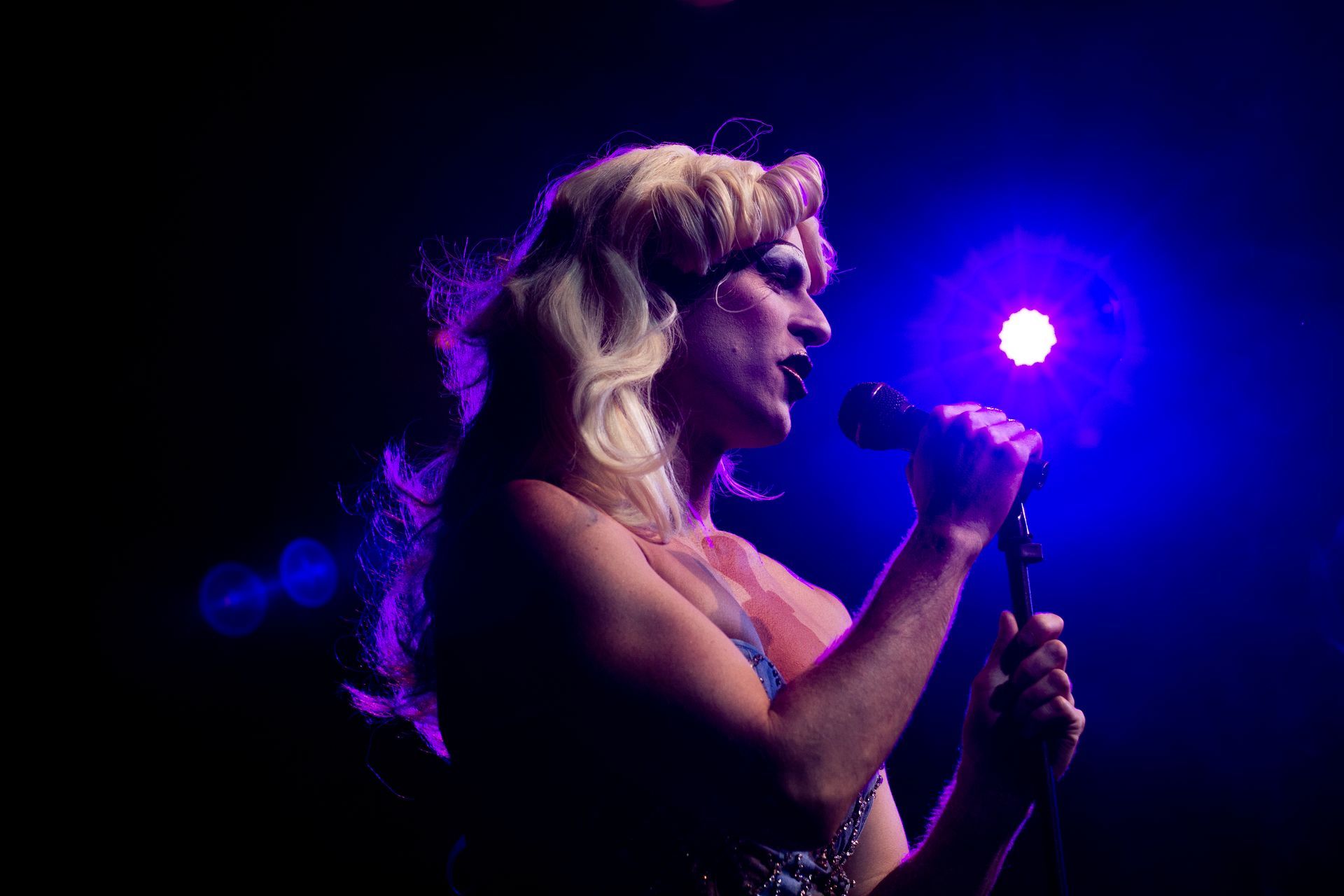 Drag performer with blonde wig sings into a microphone, bathed in blue stage light.