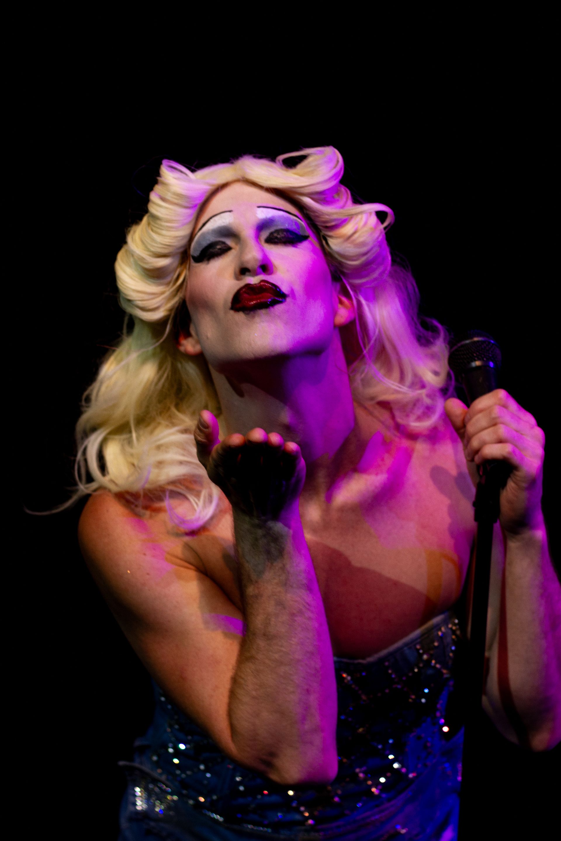 A drag queen in a blonde wig blows a kiss while holding a microphone in a stage setting.