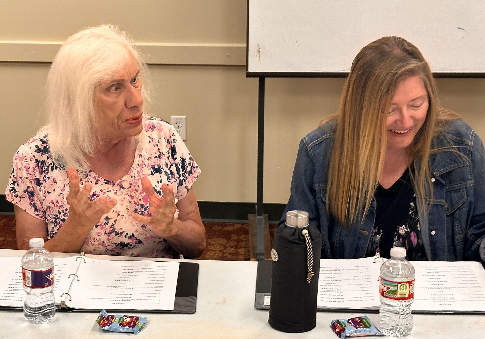 Two people seated at a table, one gesturing while the other smiles, with water bottles and papers in front of them.