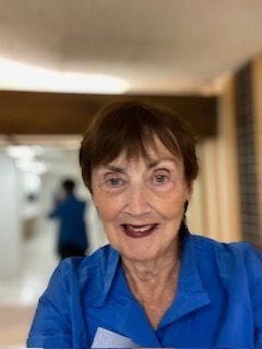 Elderly woman with short brown hair smiles at camera, wearing blue collared shirt. Background blurred.