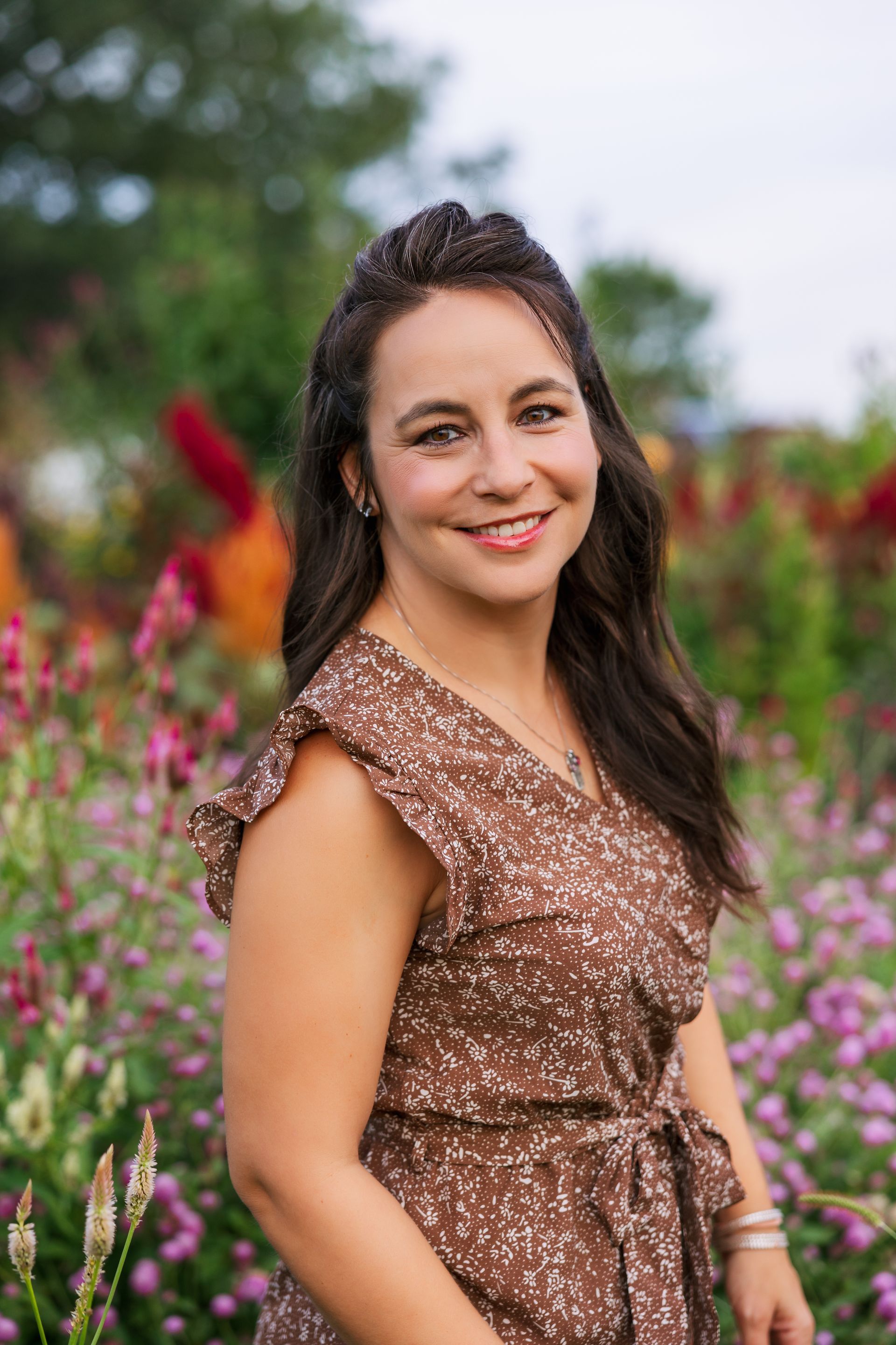 Woman smiling in a brown dress standing in a flower garden with pink and red blooms.