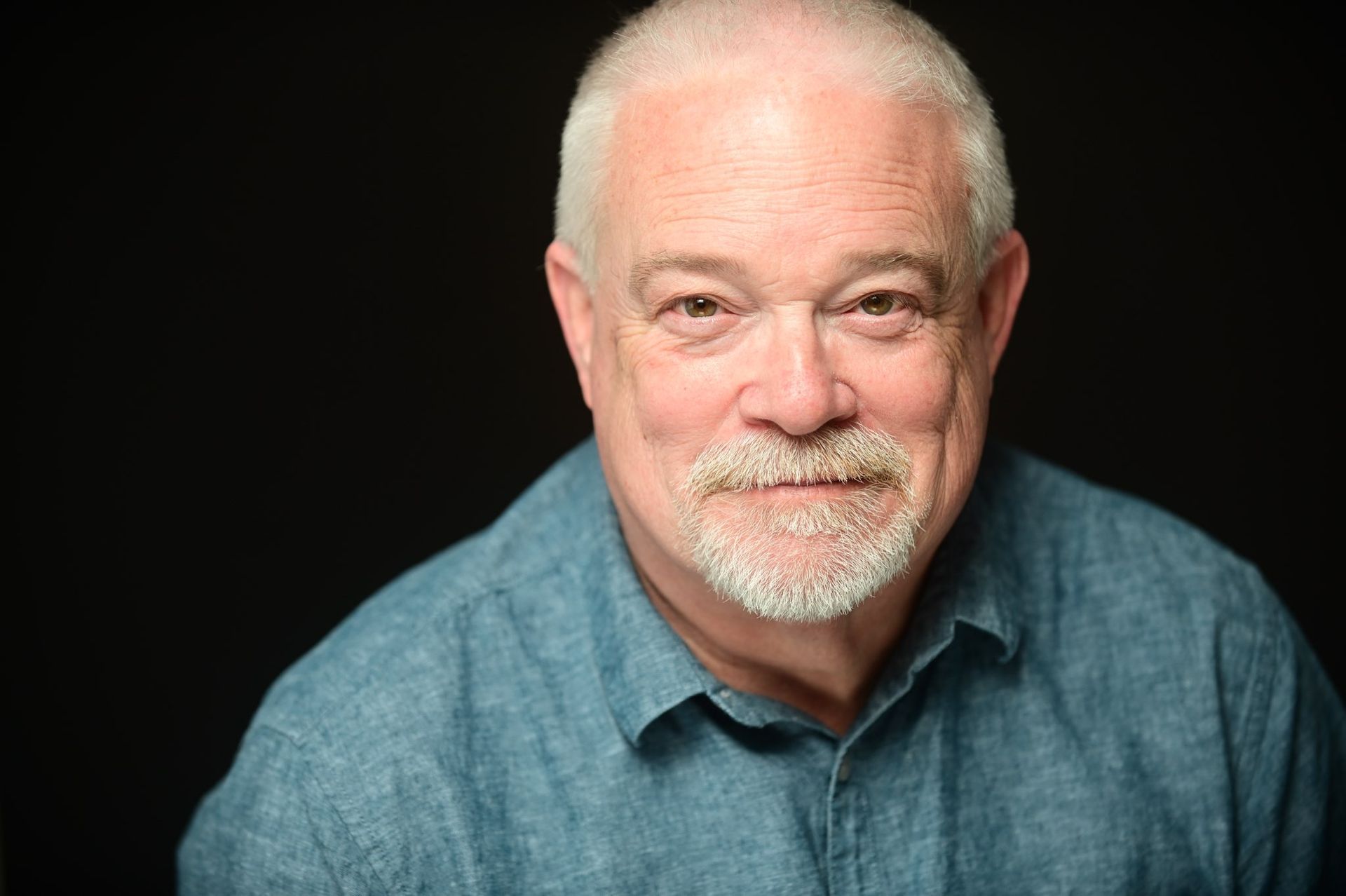 Smiling white-haired man in a blue shirt against a dark background